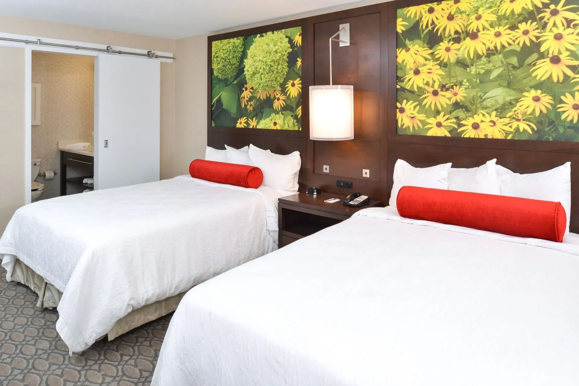 A hotel room with two queen beds featuring white bedding, red bolster pillows, and floral wall art above the headboards.