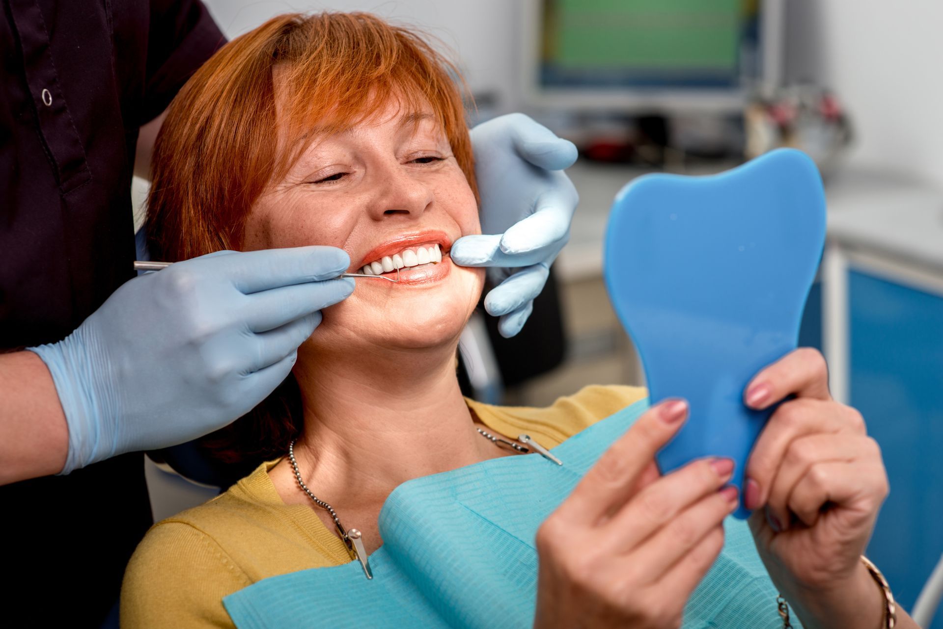 A woman is sitting in a dental chair looking at her teeth in a mirror.