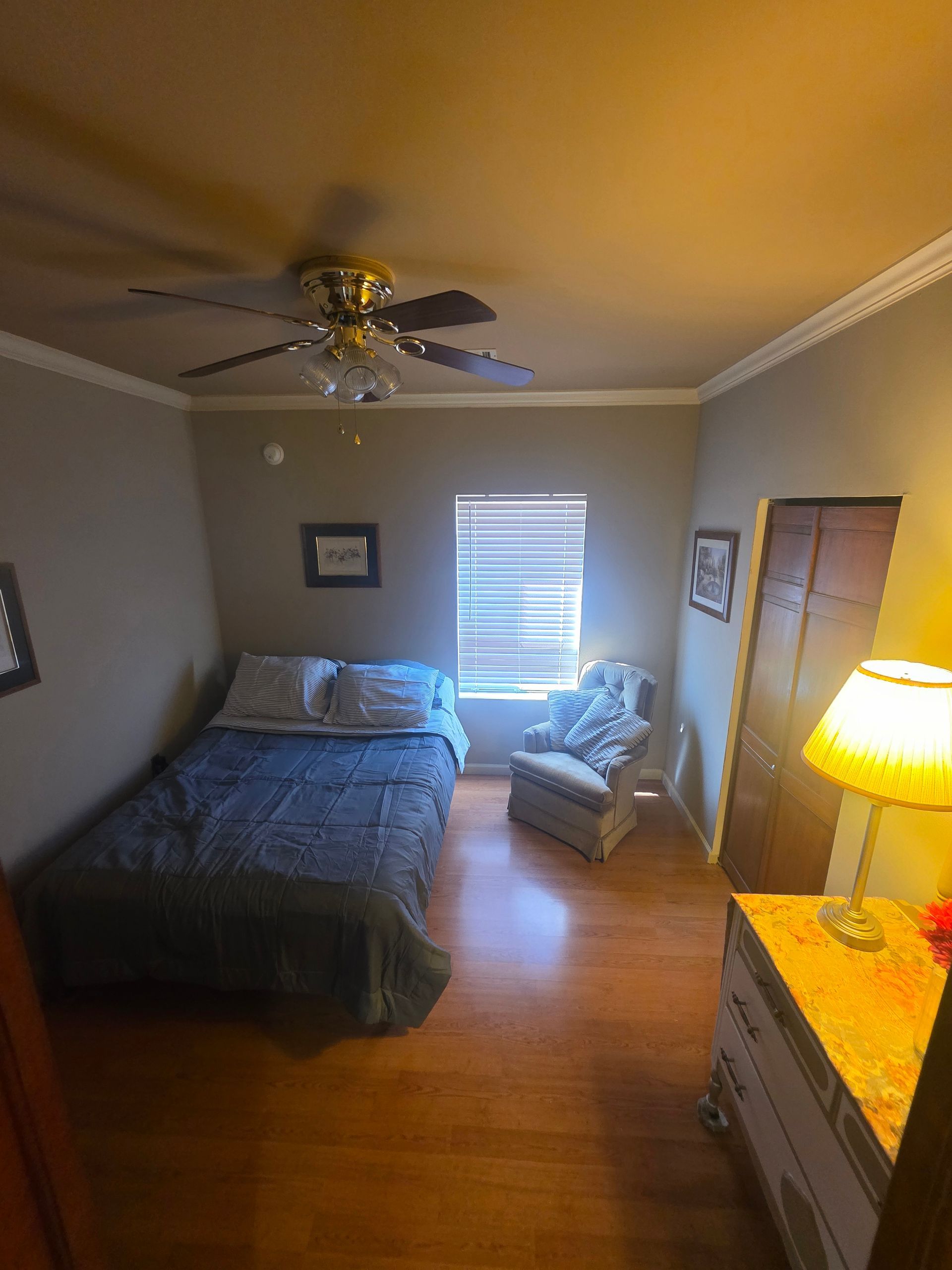 Bedroom with a bed, chair, dresser, and wooden floor. Neutral walls and ceiling fan.