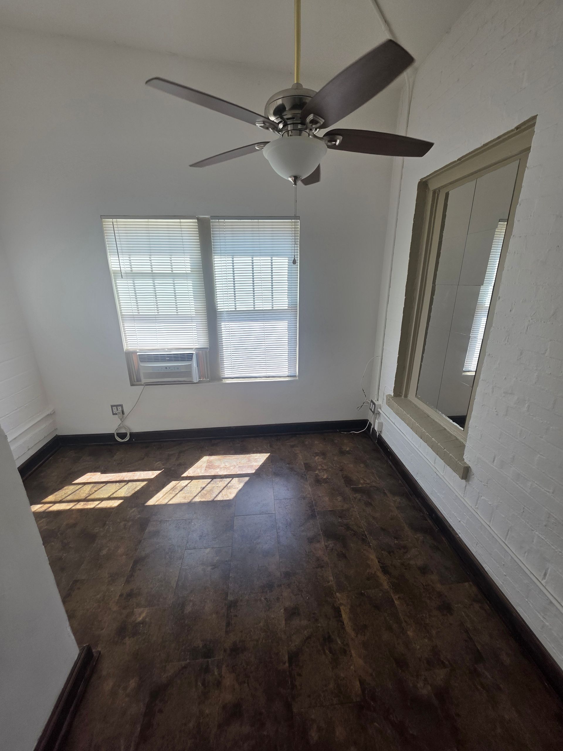 Empty room with hardwood floors, window, mirror, ceiling fan, and white walls.