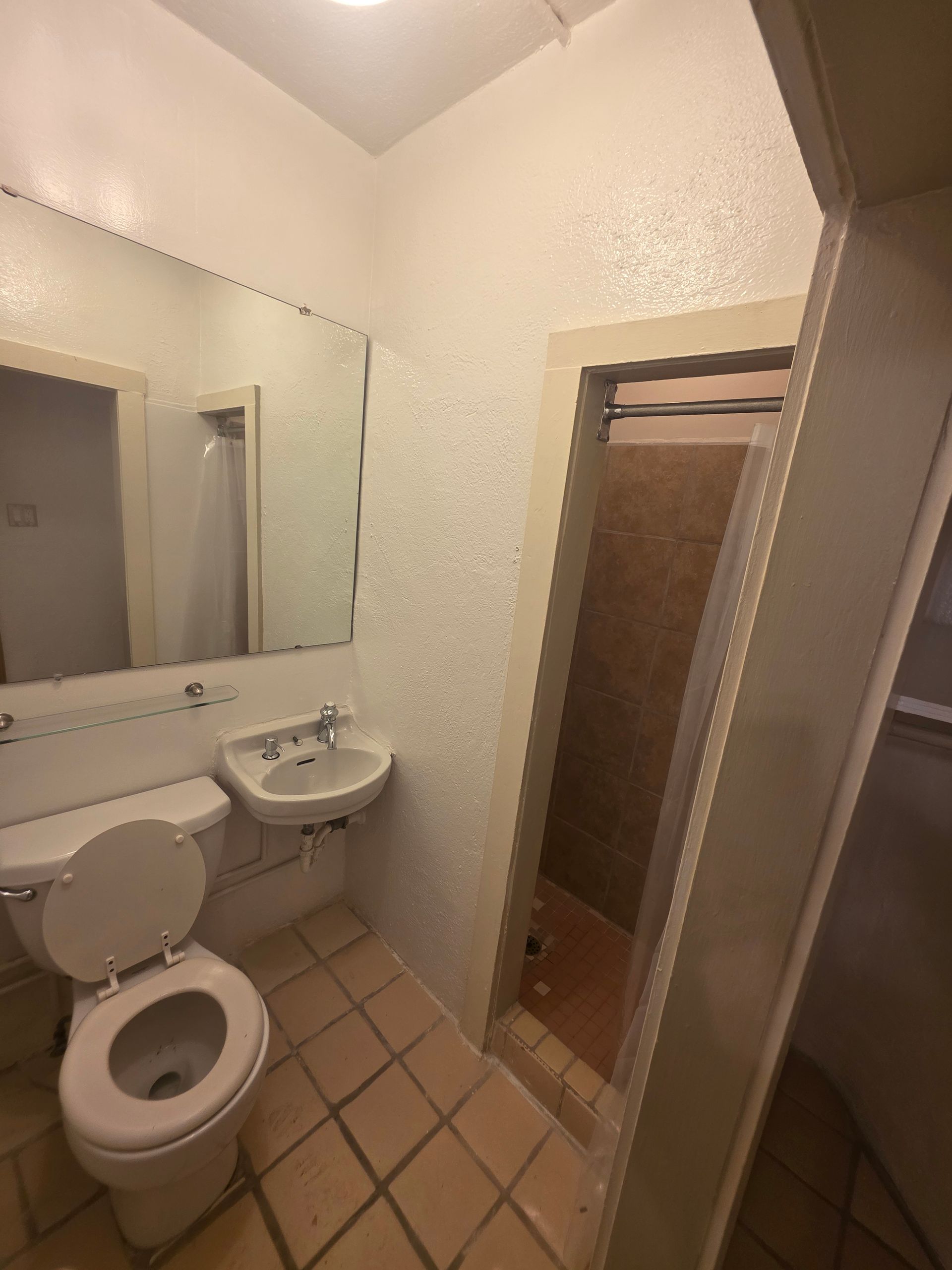 Small bathroom with toilet, sink, mirror, and shower with sliding door. Beige tile and white walls.