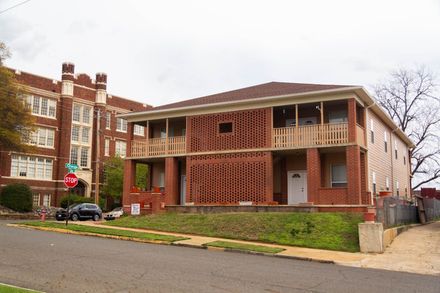 Two-story brick building with balconies next to a large red brick building. A street sign and car are in the foreground.