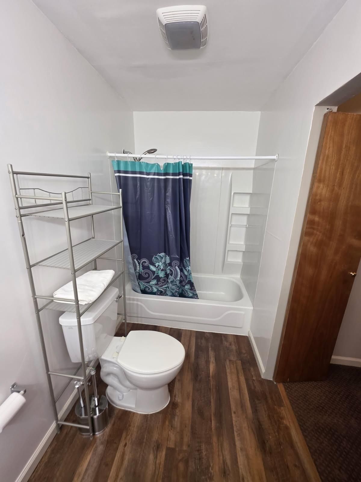 Bathroom with toilet, tub/shower, dark wood floor, and a metal shelf unit.