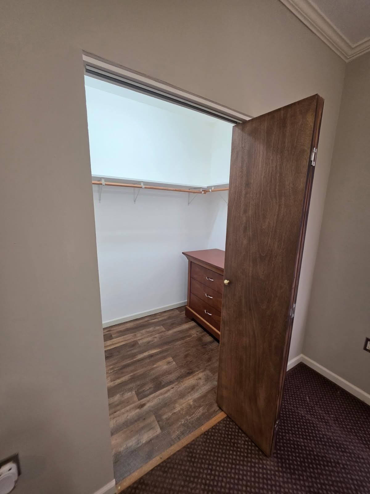 A walk-in closet with a wooden door ajar. The interior has shelves, a dresser, and wood-look flooring.