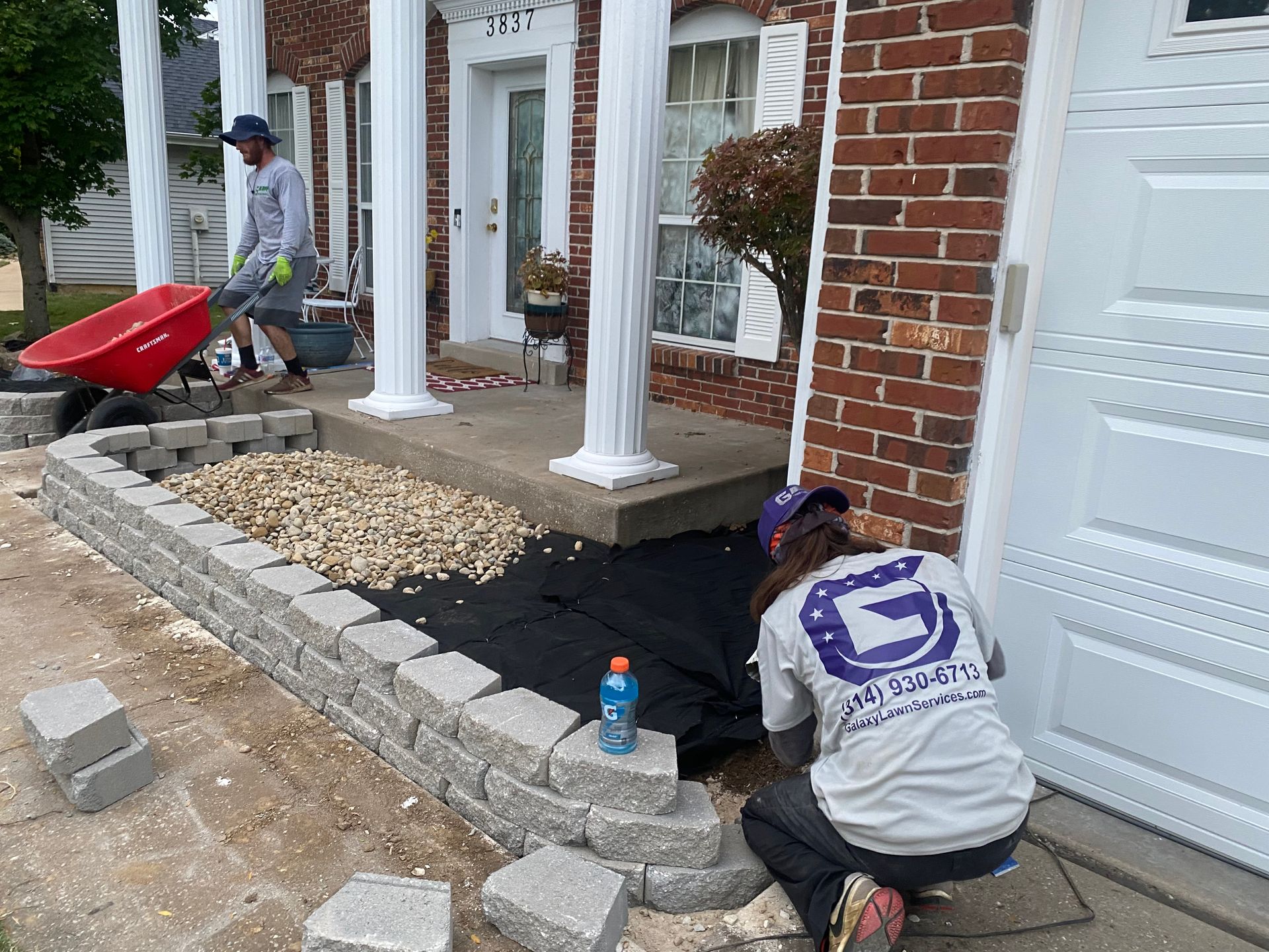 Workers constructing a small retaining wall in front of a brick house with columns.