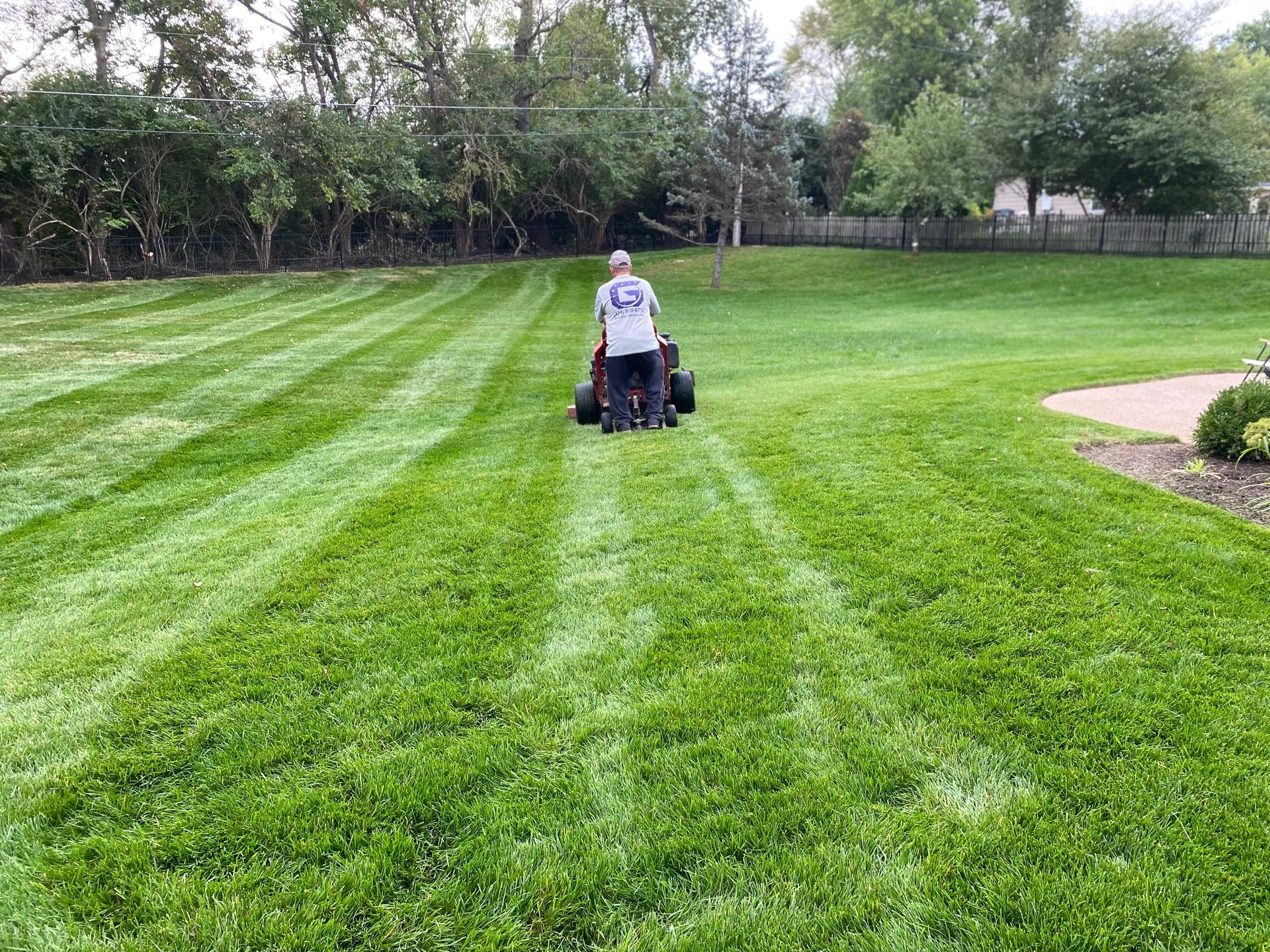Person mowing a large, green lawn, creating striped patterns. Outdoors, trees in background.
