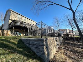A two-story house with a wooden deck and a black metal fence on a retaining wall, under a clear blue sky.