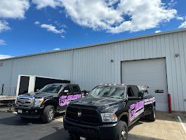 Two black pickup trucks with purple logos parked outside a white industrial building under a cloudy blue sky.