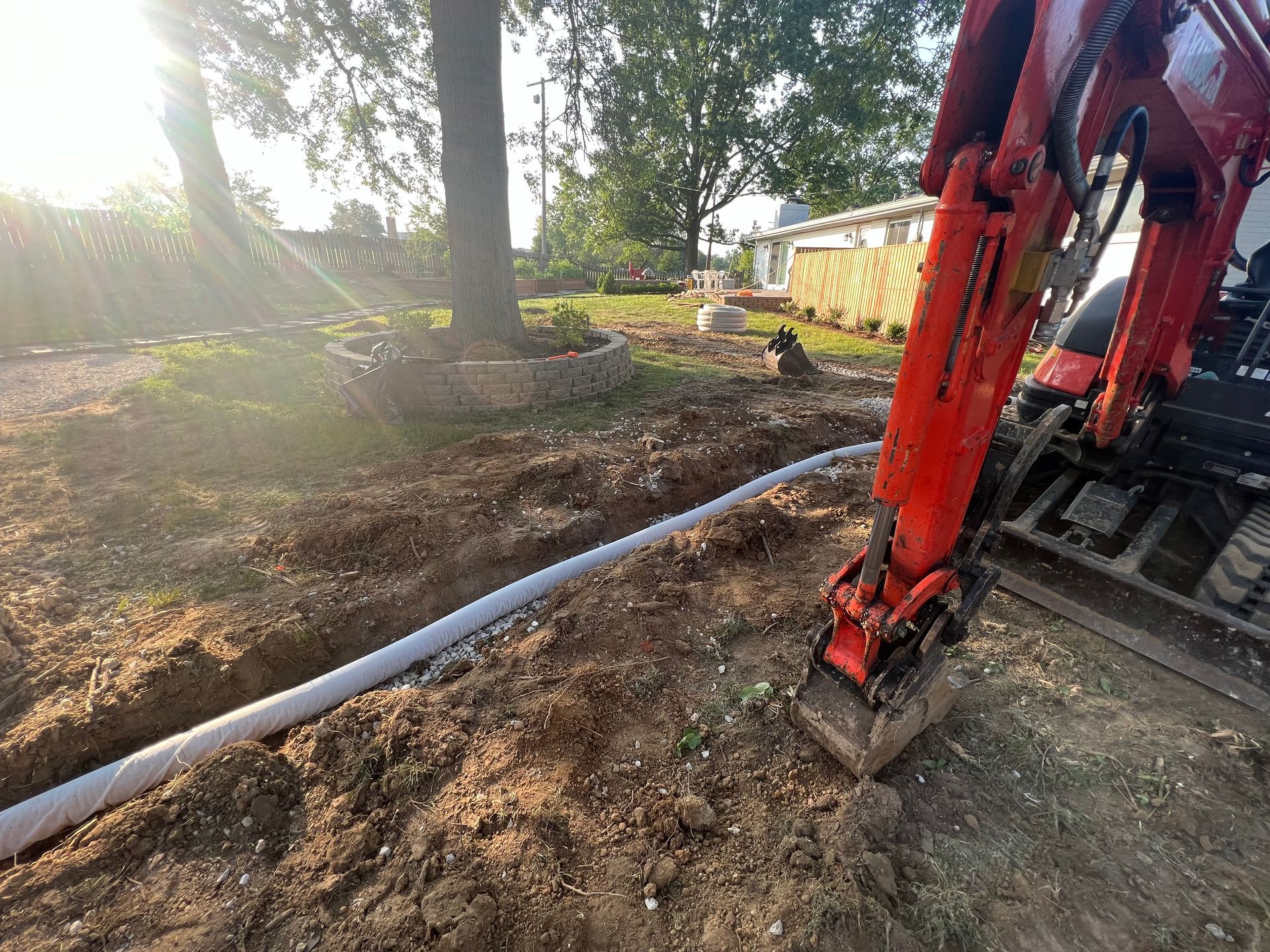 An excavator digs a trench in a yard, placing a white drainage pipe.