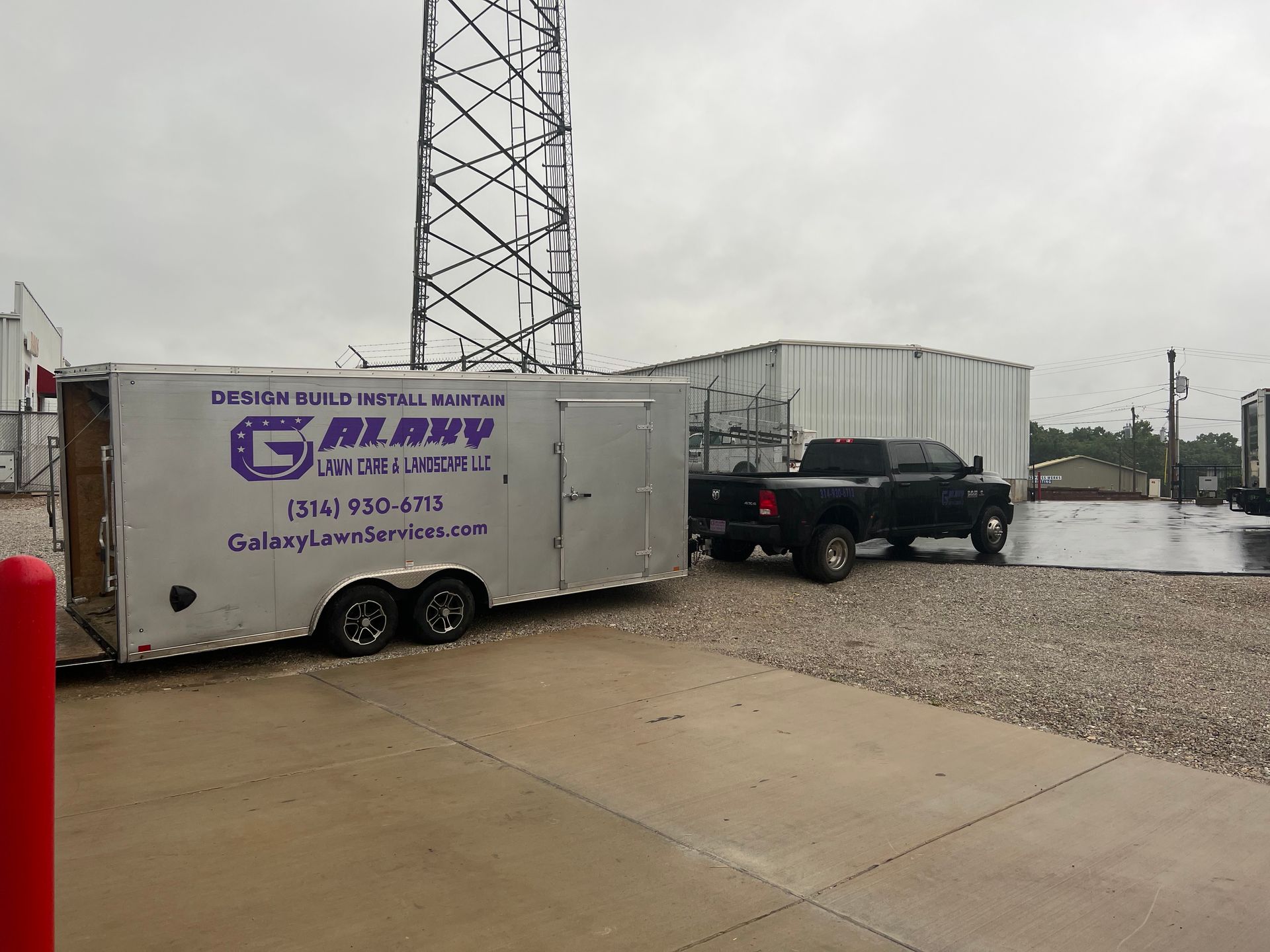 A silver trailer with company logo attached to a black truck, parked near a cell tower and container.