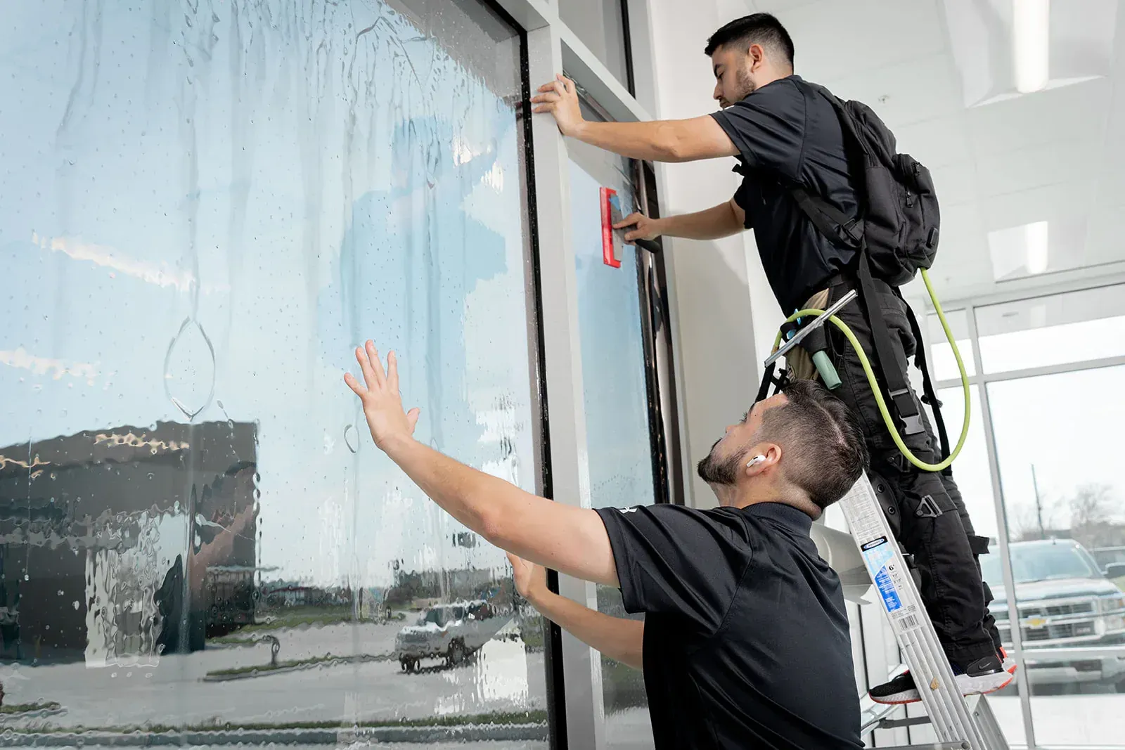 Two men installing window tint on a storefront. One on a ladder, squeegeeing, while the other smooths the film.