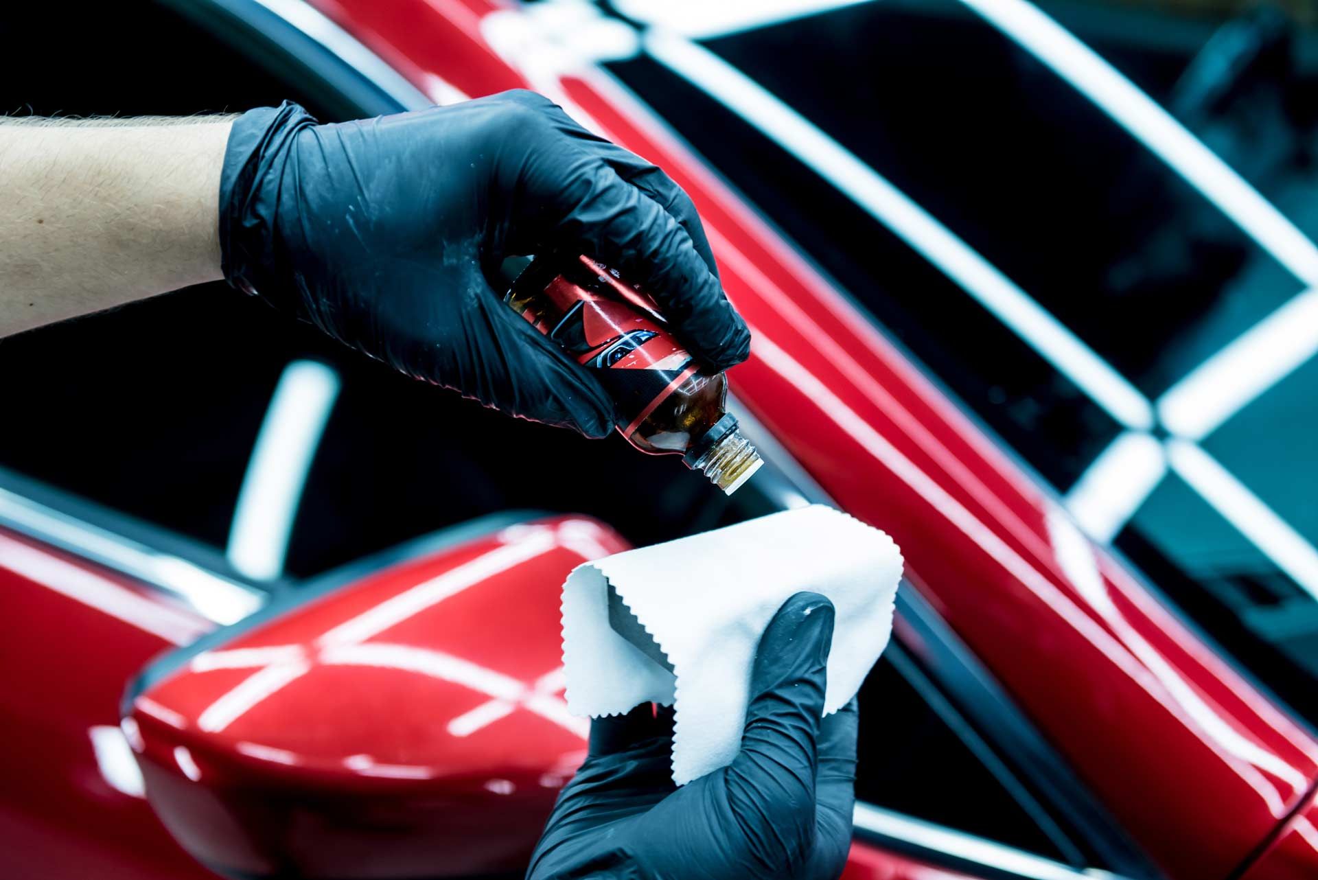 Gloved hands applying car ceramic coating to a white applicator pad near a red car mirror.