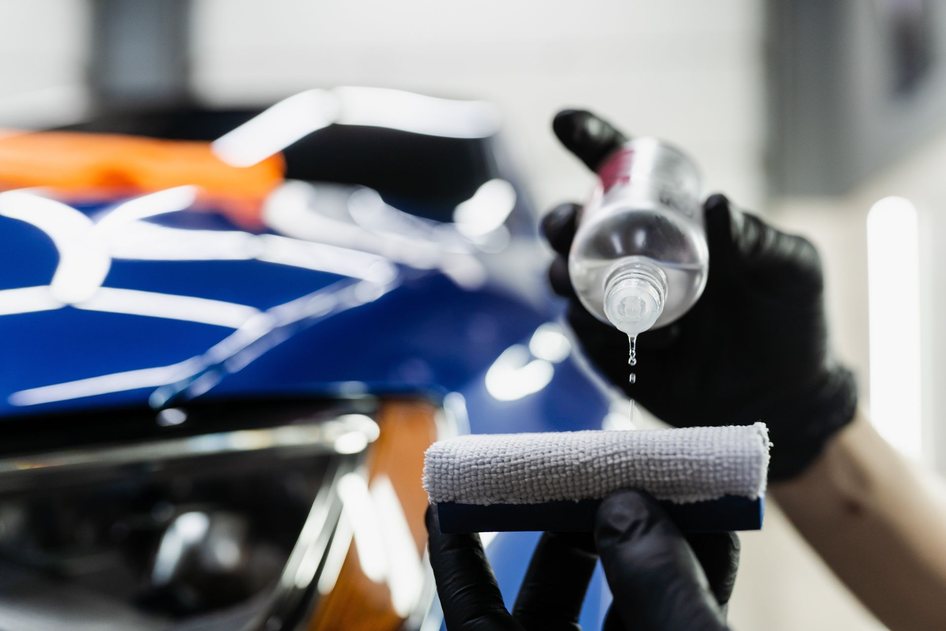 Person wearing black gloves applying liquid onto a microfiber cloth next to a blue car.