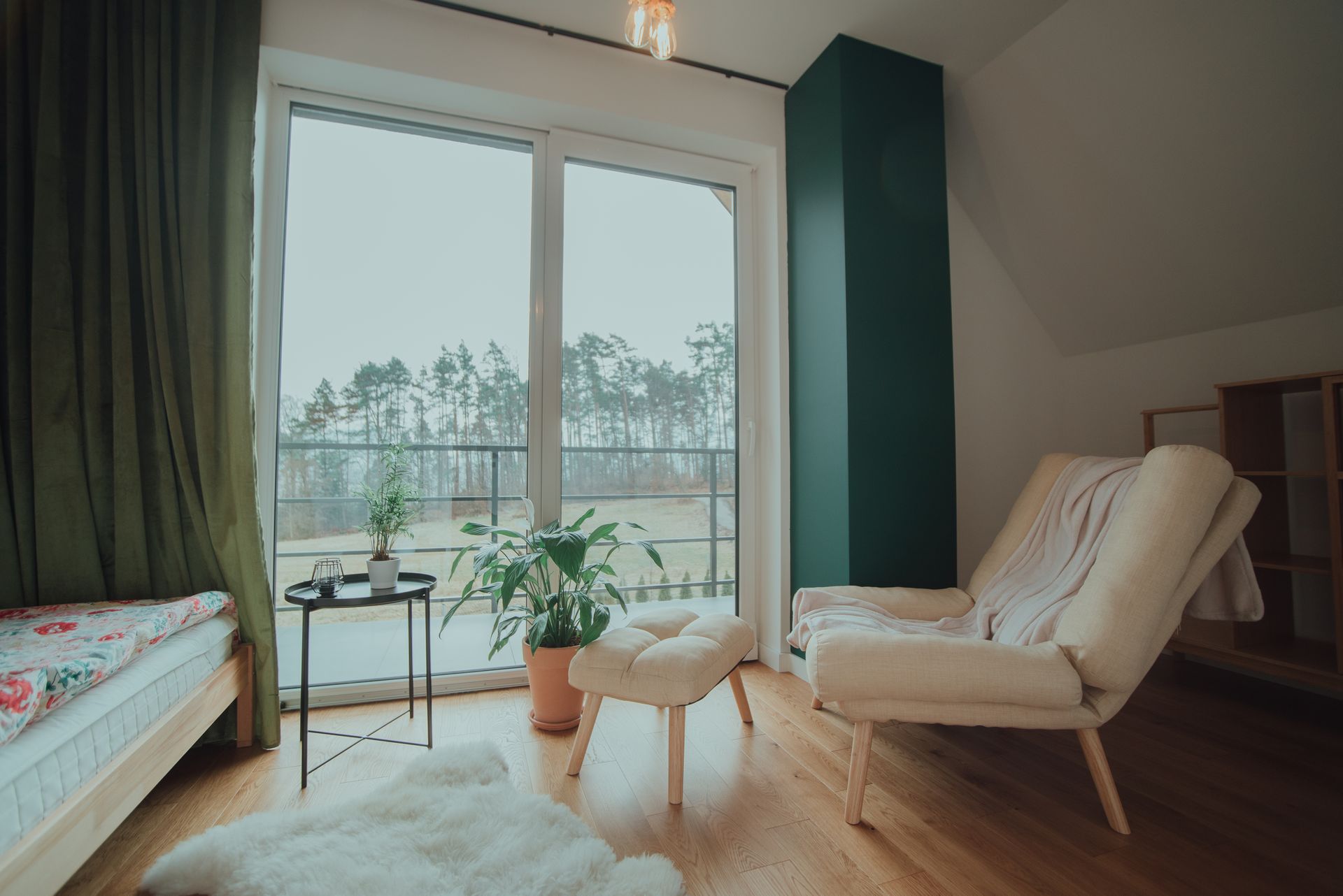 Cozy room with large window overlooking a forest. Cream-colored chair and ottoman, green curtain, bed, and plants.