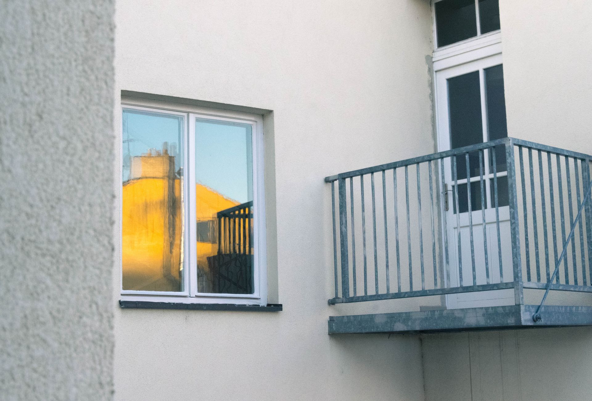 Window reflecting a yellow building; balcony with metal railing and white door on light-colored building.