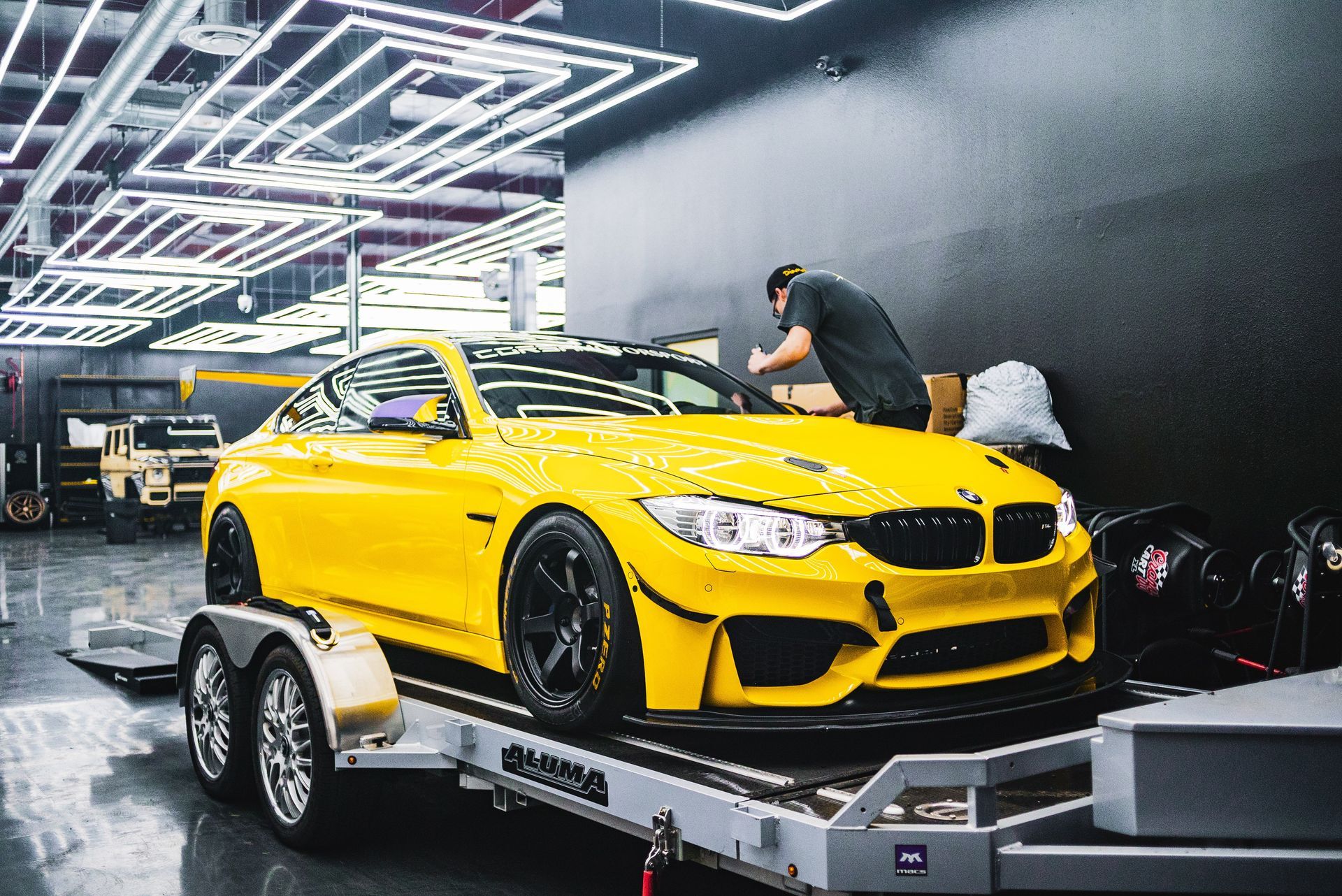 Yellow BMW M4 on a trailer inside a garage, mechanic working on it.