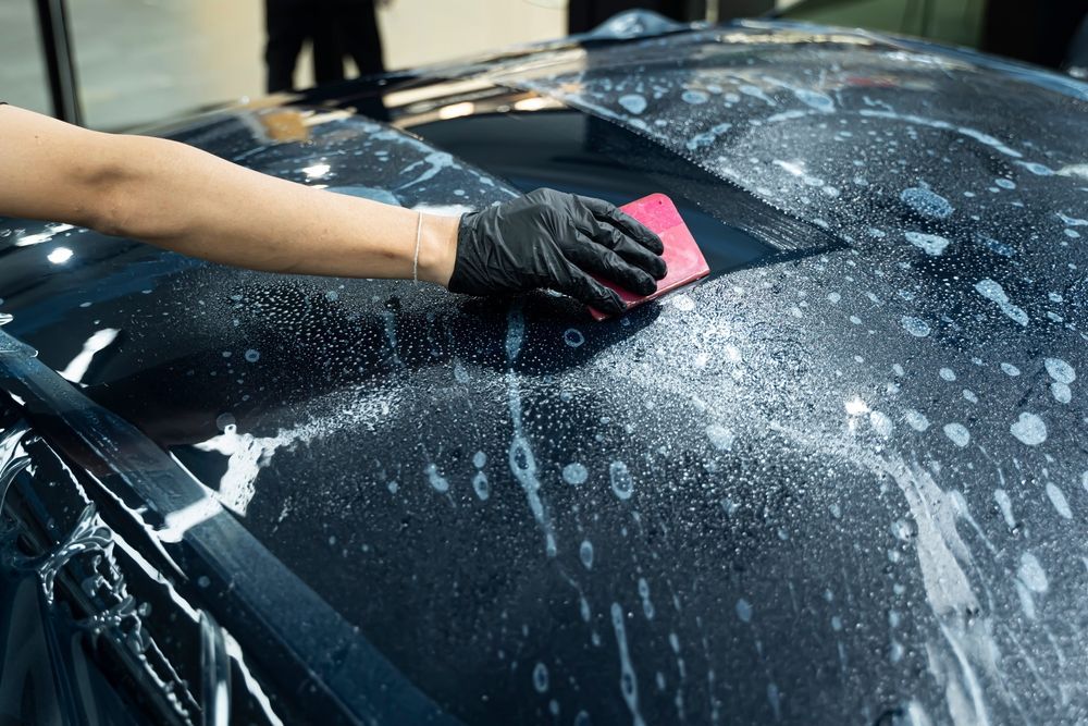 Person applying film to a car's hood, using a squeegee. Black gloves, soapy water visible.