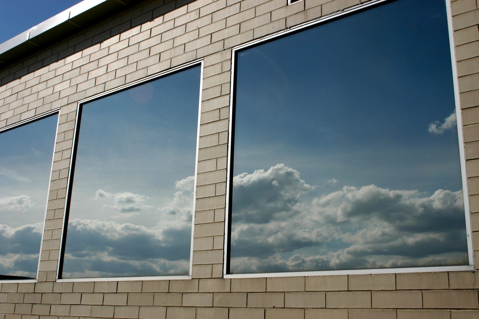 Three windows reflecting a cloudy blue sky on a beige brick building.