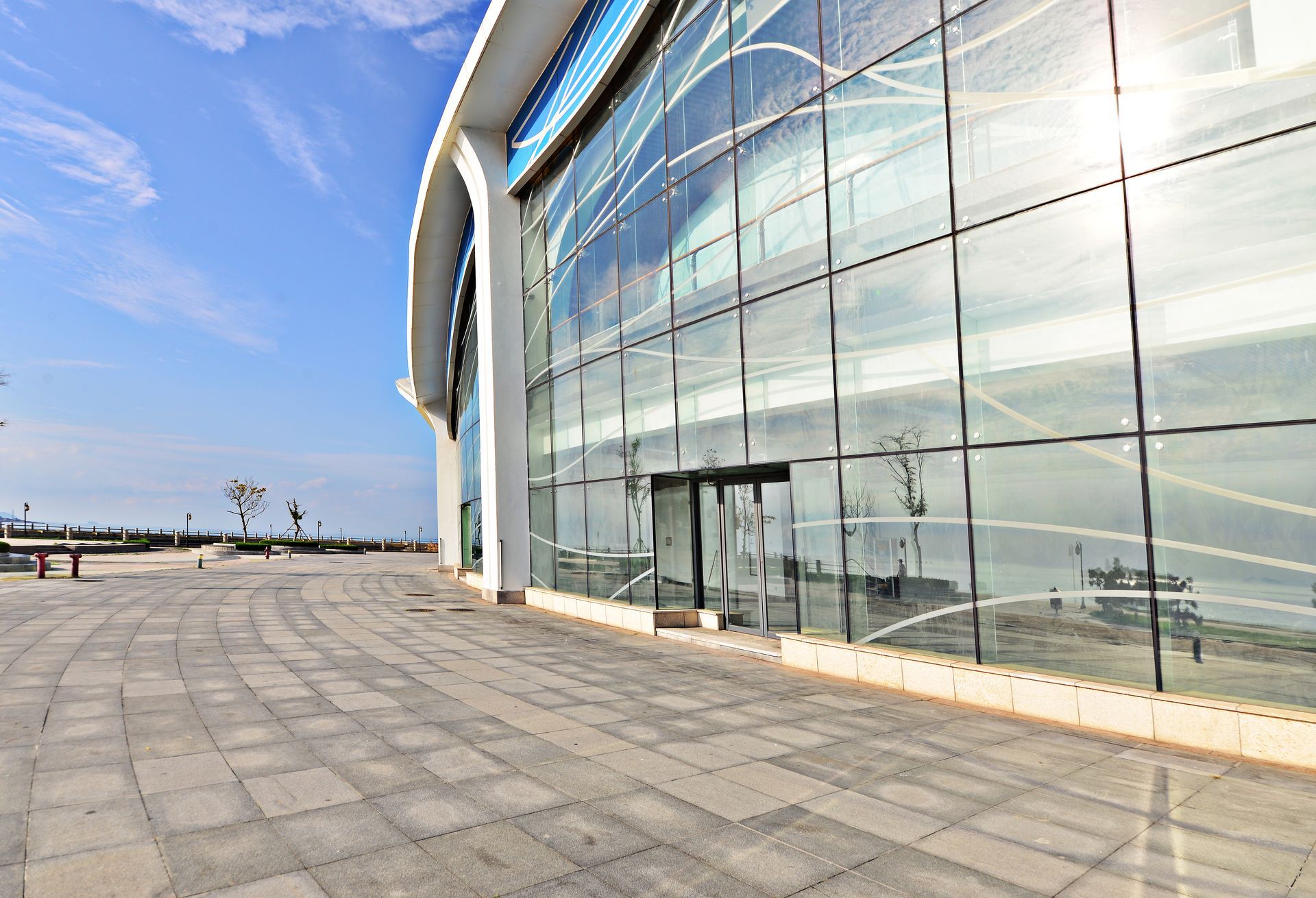 A large glass-walled building with a curved design, next to a paved walkway under a blue sky.