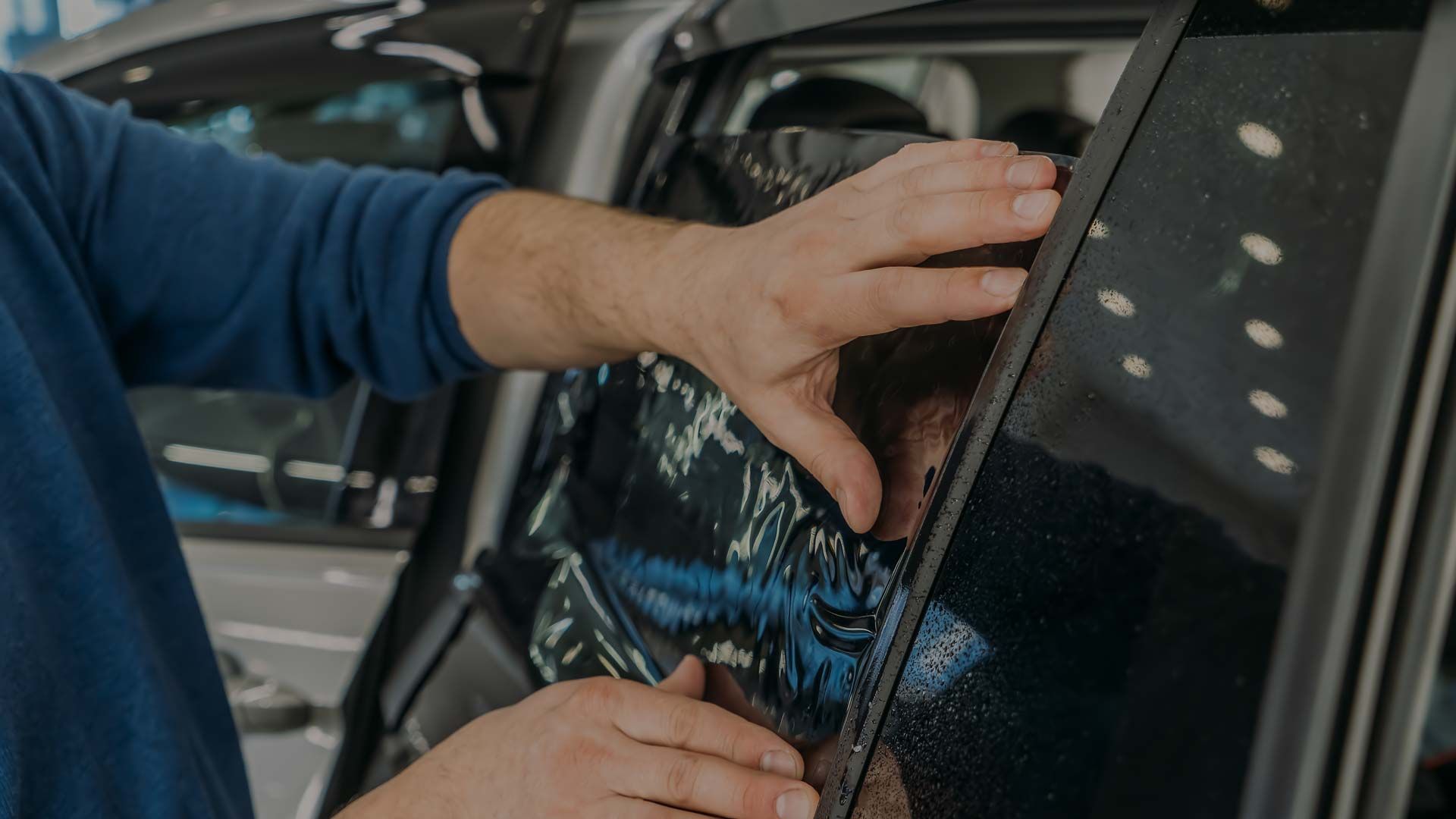 Hands applying window tint to a car window in a garage.