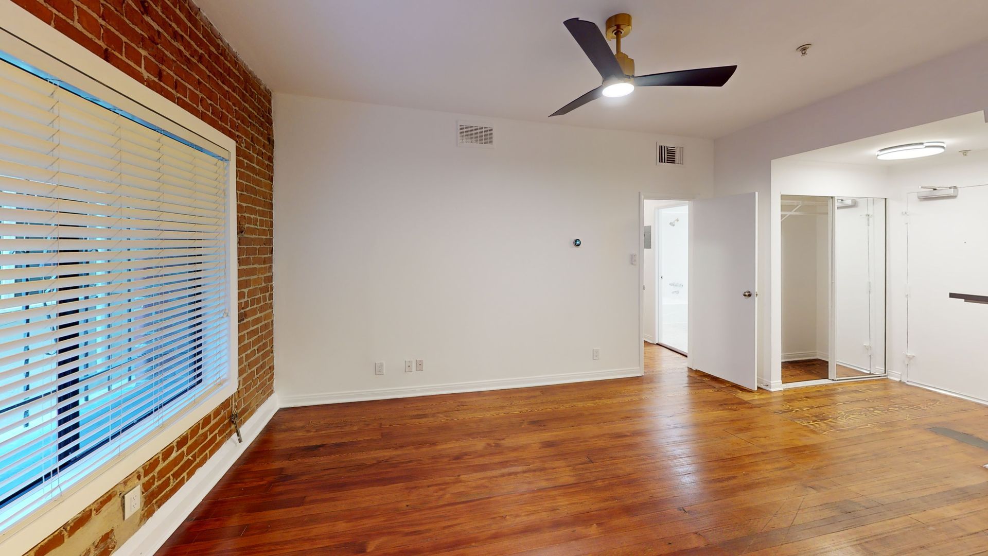 Empty apartment interior with exposed brick, wood floor, and a ceiling fan.