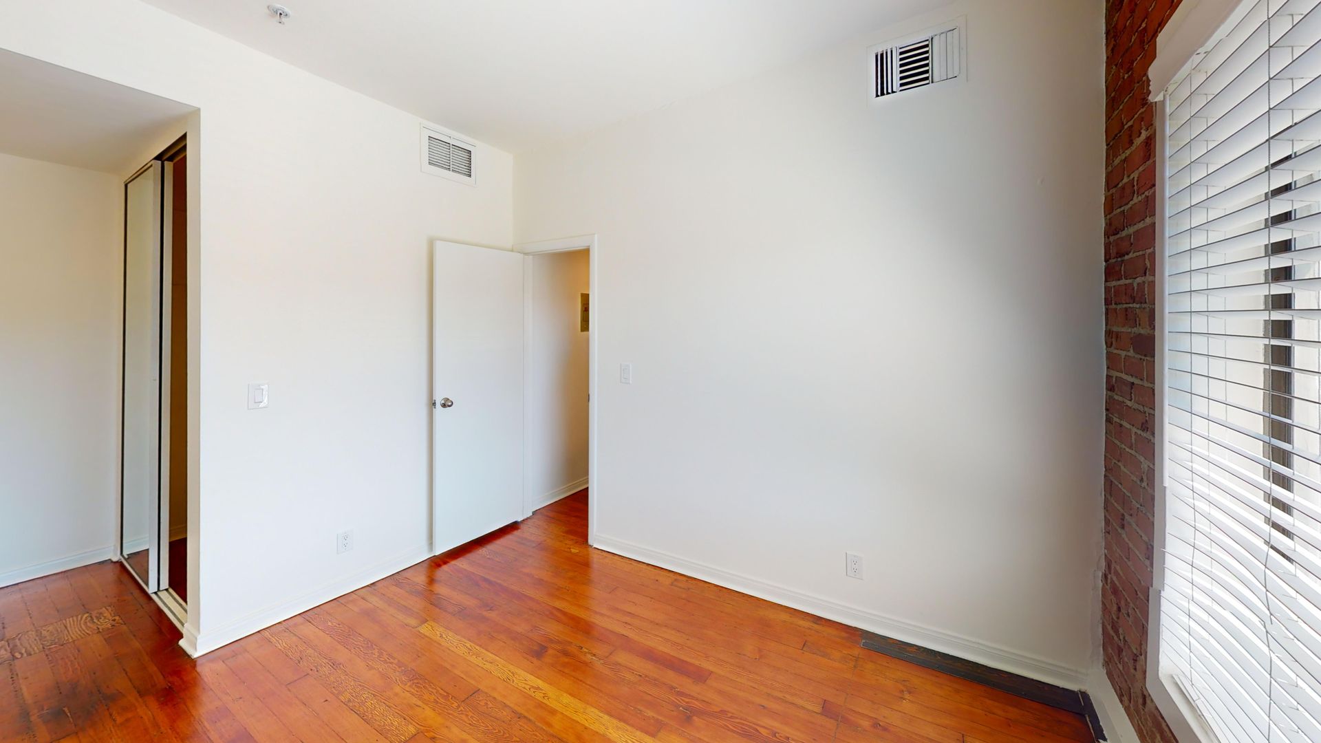 Empty bedroom with hardwood floors, white walls, closet, door, and window with blinds.