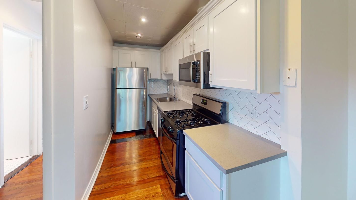 Narrow kitchen with stainless steel appliances, white cabinets, light gray countertops, and wood floor.