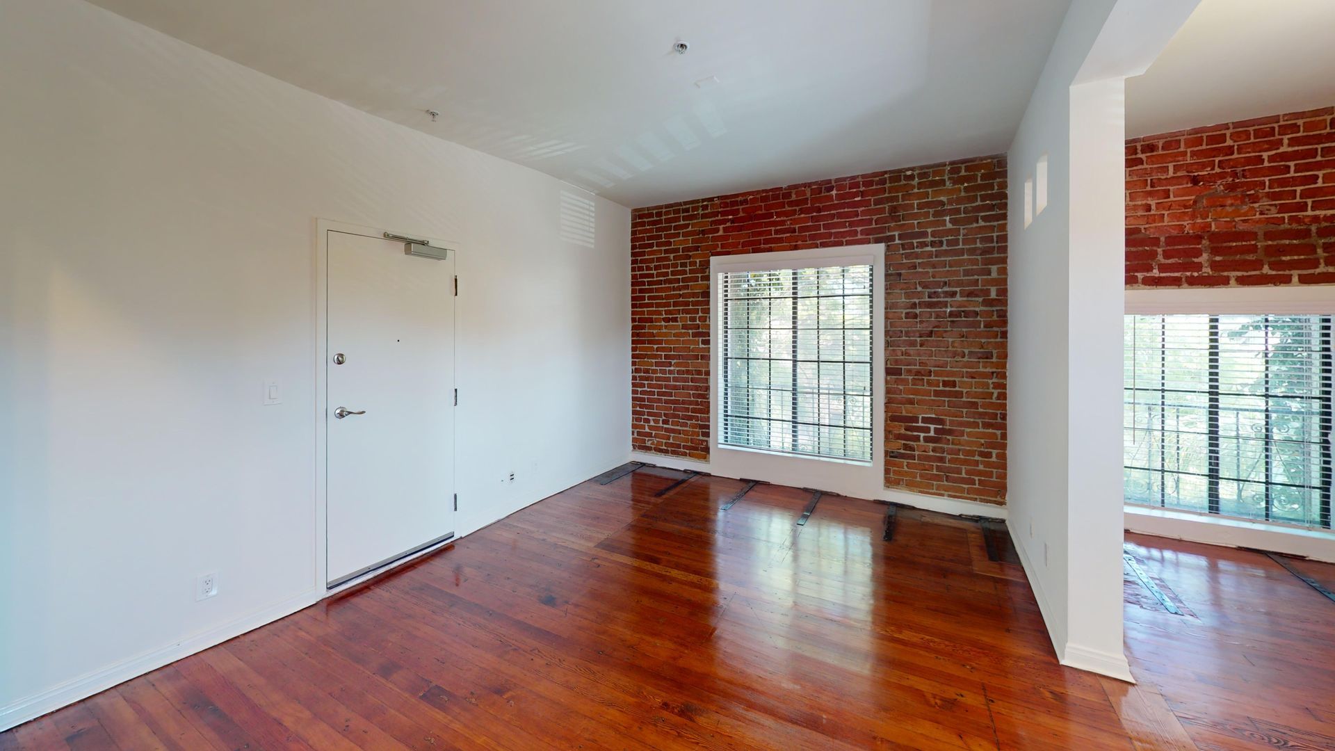 Empty room with red brick wall, windows, and wood flooring. White walls and door.