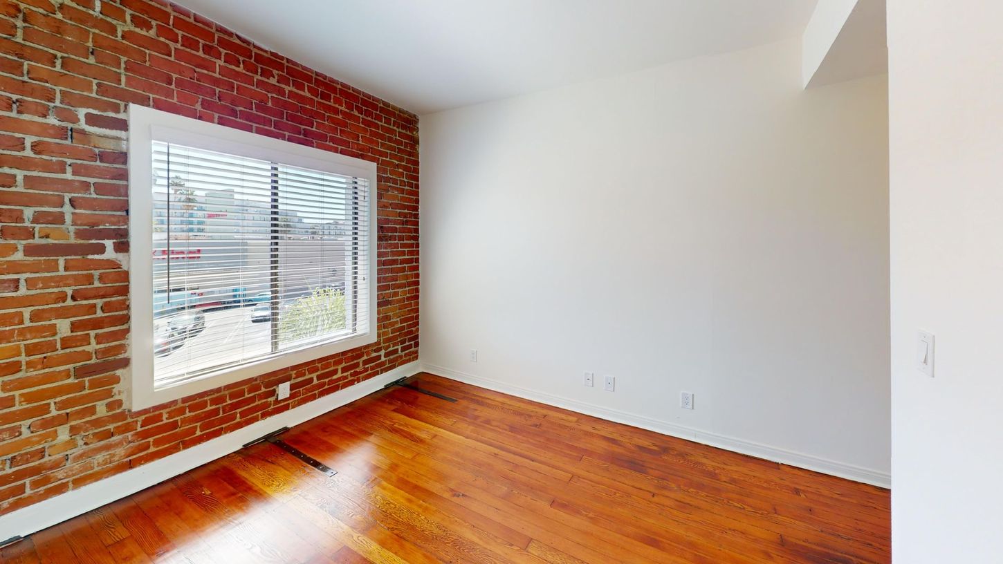 Room with exposed brick wall, window with blinds, and hardwood floors.