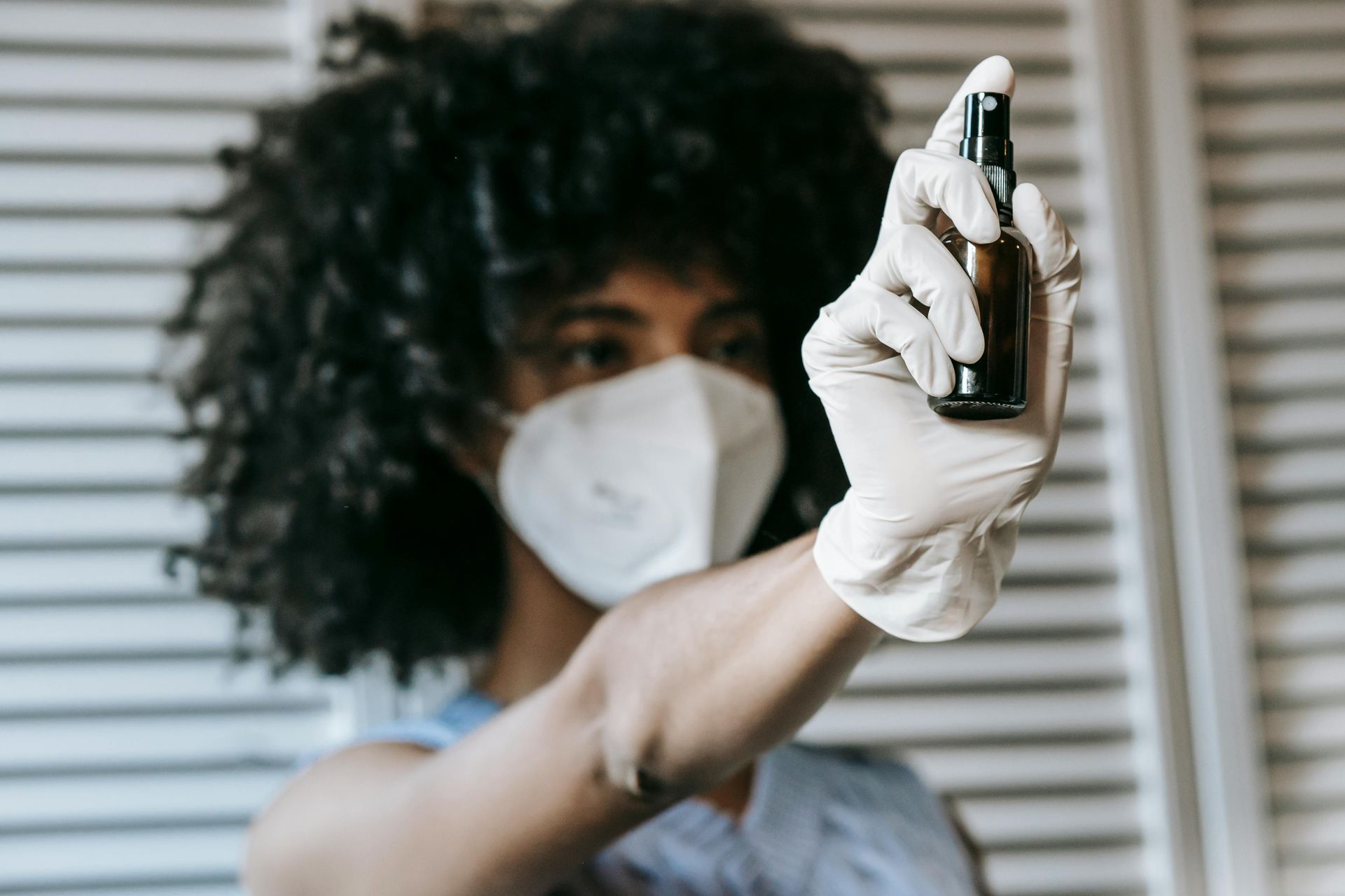 Woman wearing mask and gloves, holding a spray bottle in front of a white shutter.