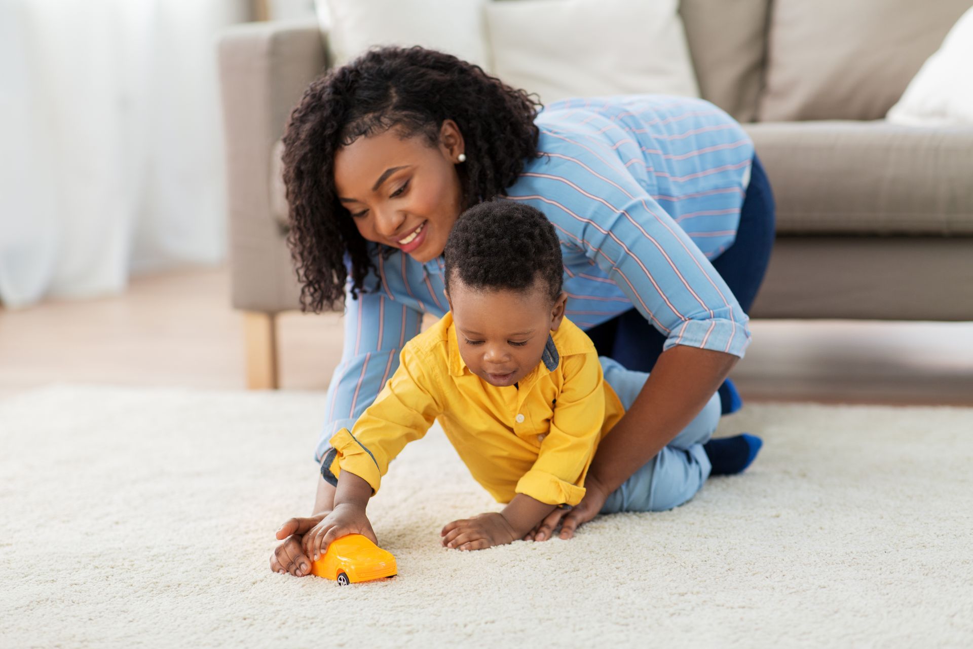 Woman and baby playing with a toy car on a rug. The woman kneels, smiling.