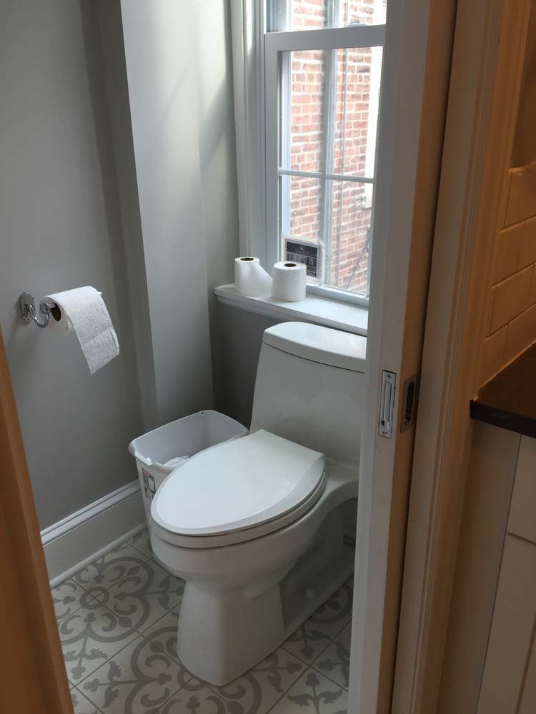 A bathroom with a toilet , trash can , and window.