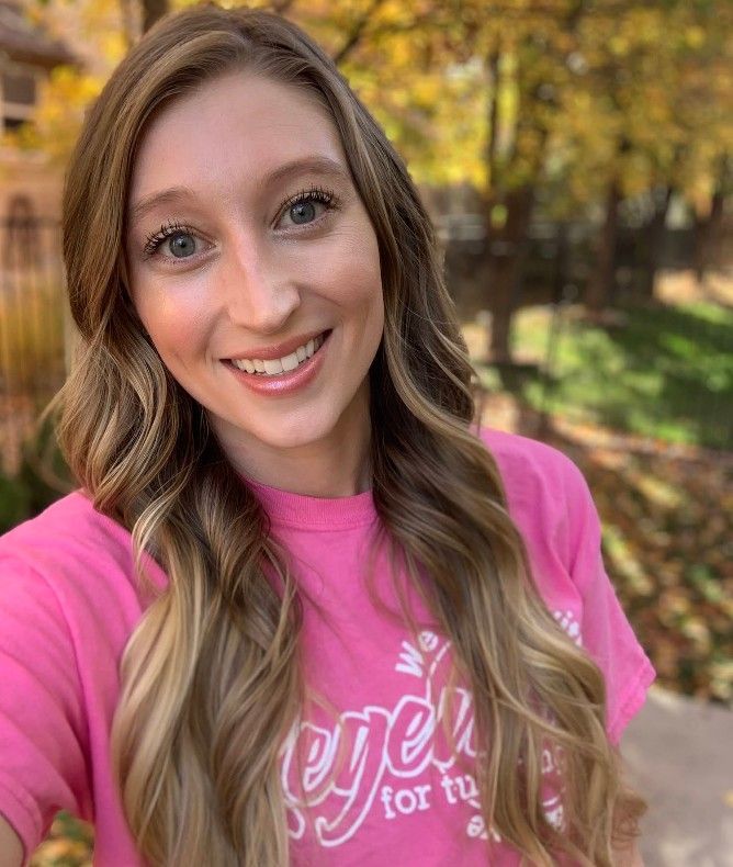 Woman with long wavy hair in pink shirt smiles outdoors in autumn setting.