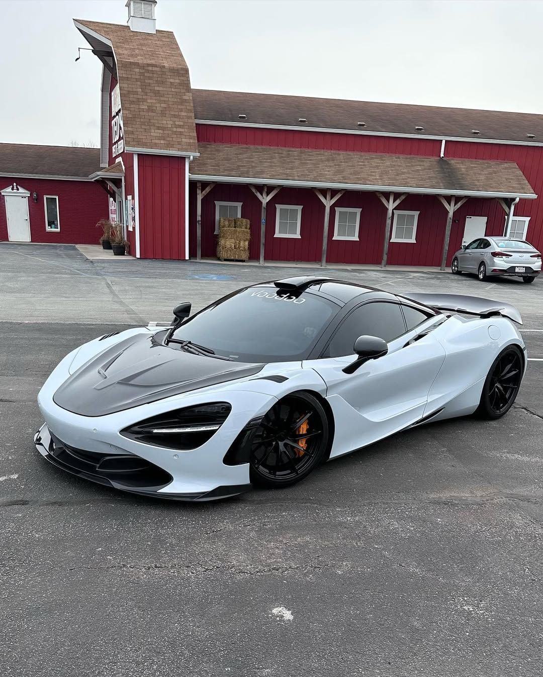 White and black McLaren 720S sports car parked in front of a red barn-like building.