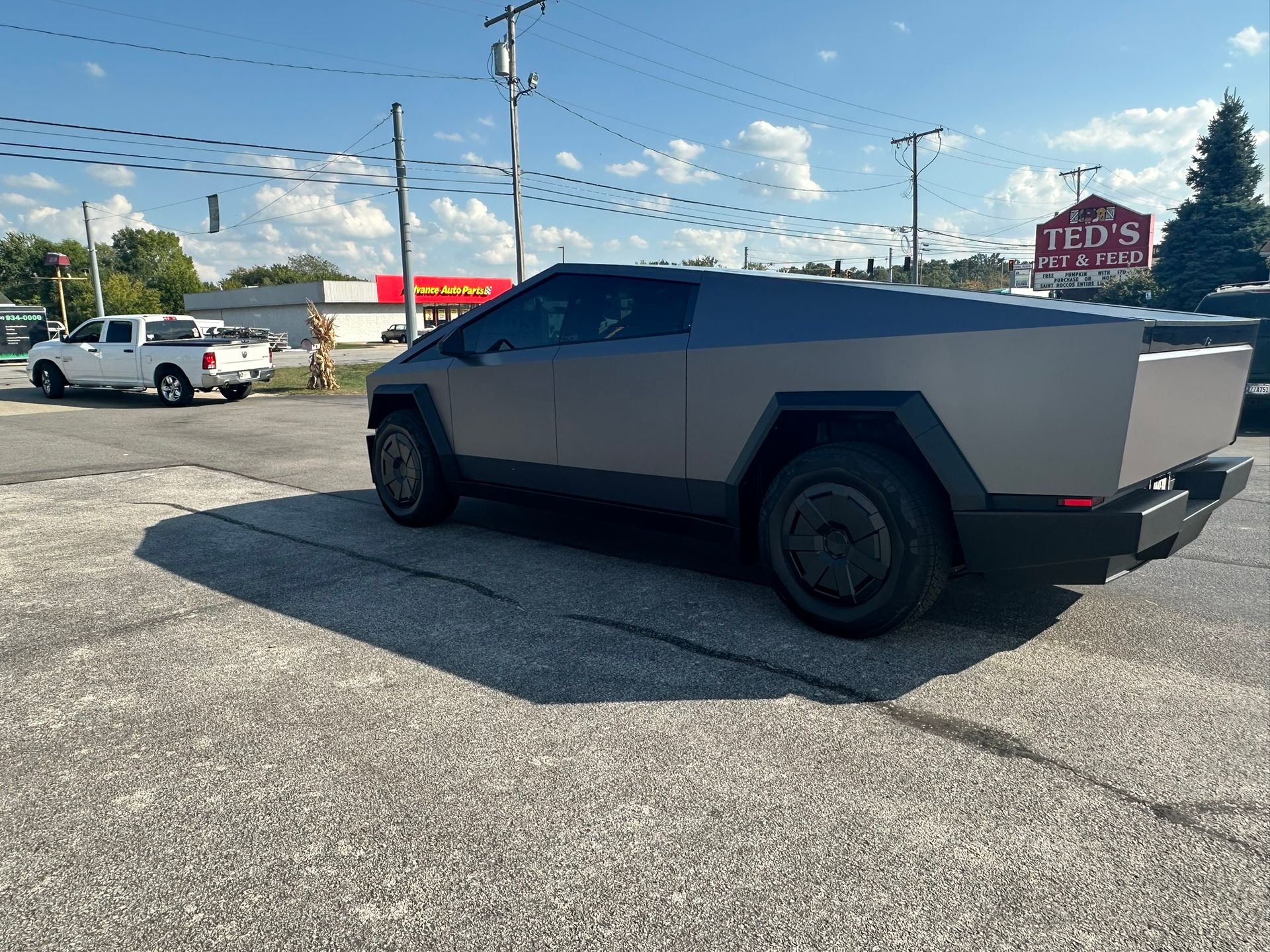 Gray Tesla Cybertruck parked on asphalt on a sunny day. A white pickup truck is in the background.