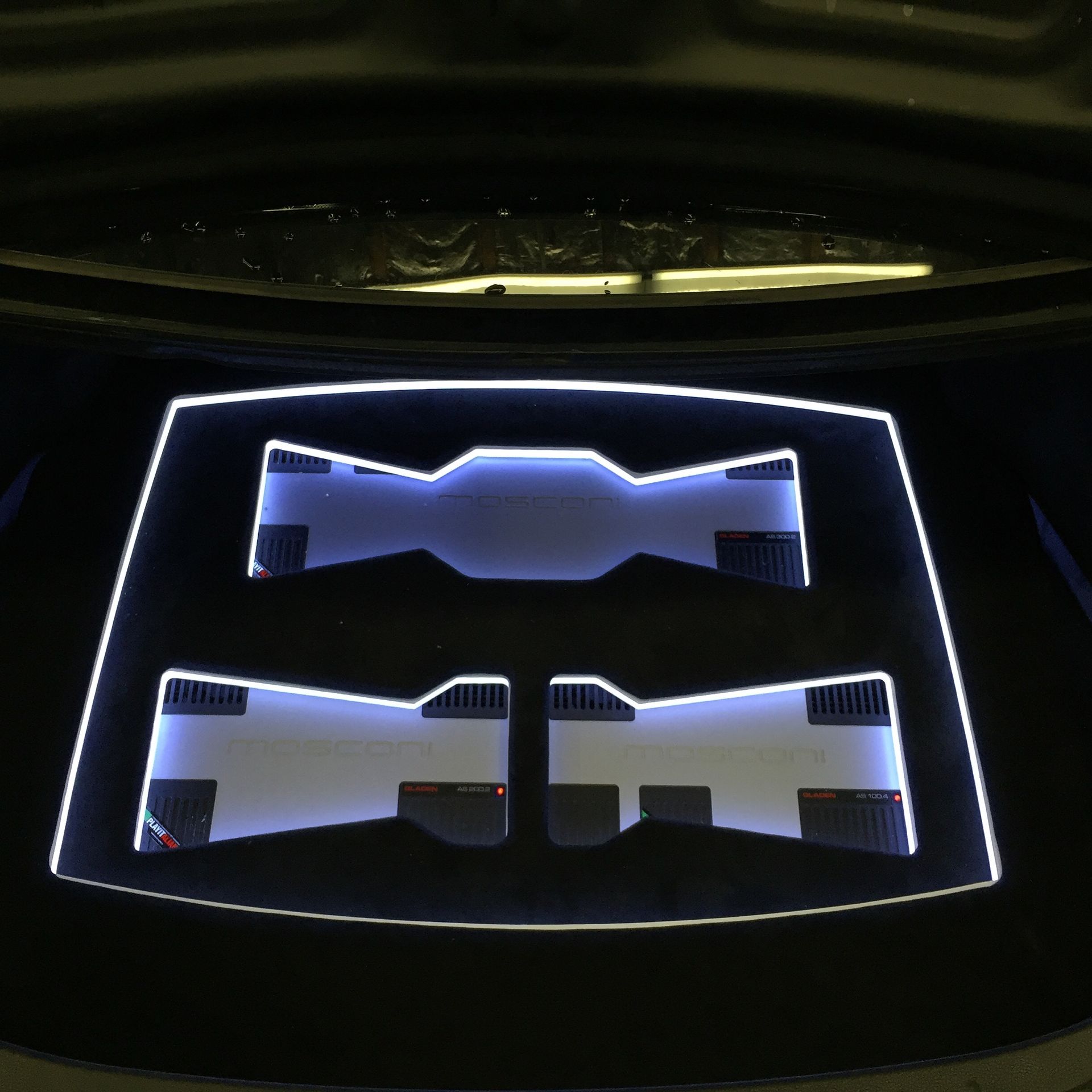 Car trunk with LED-lit panels housing audio amplifiers, glowing white and blue against a dark interior.