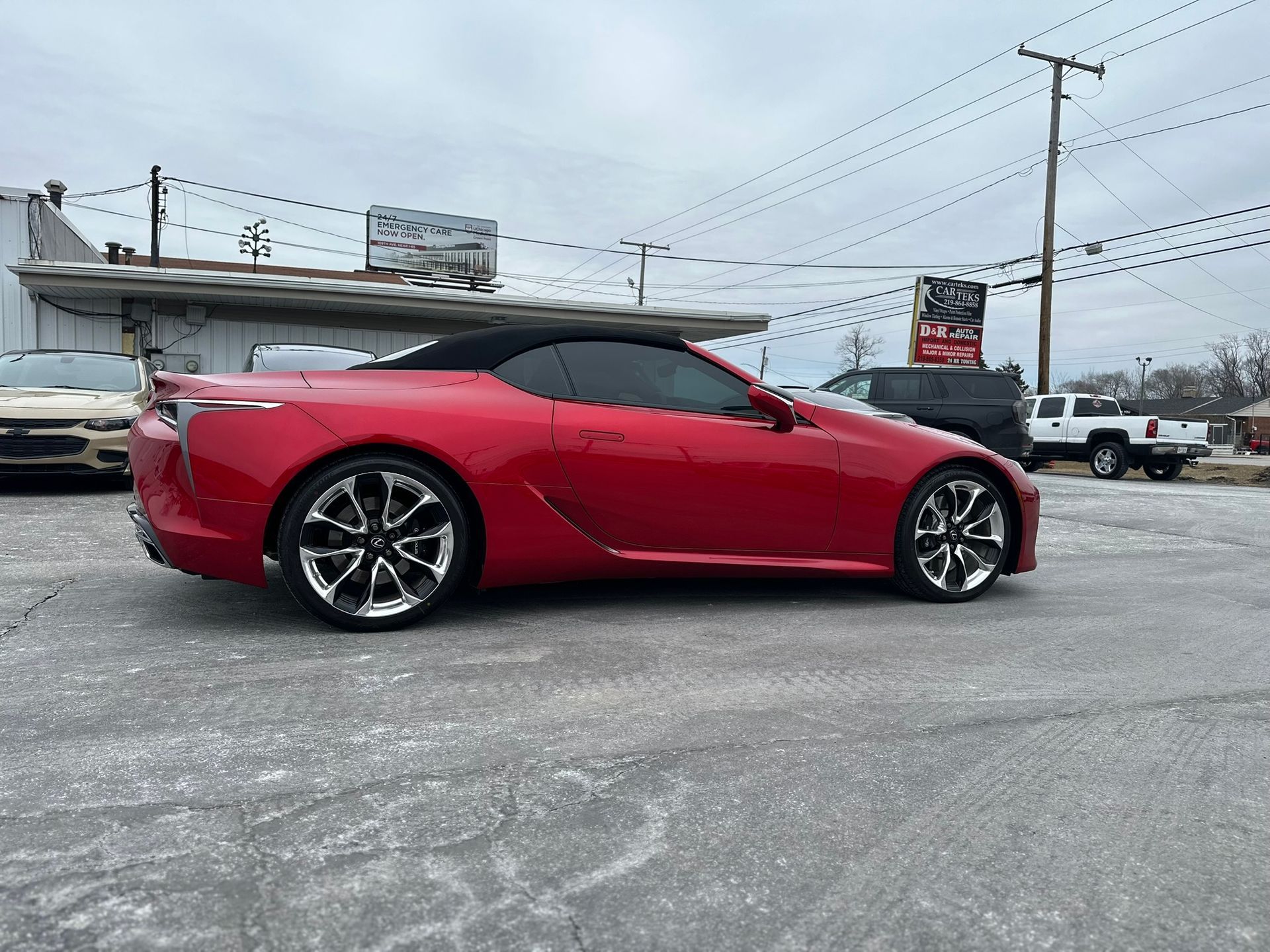 Red convertible car with black top, parked on concrete. Silver rims, cloudy sky.