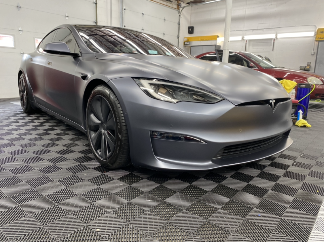 Matte gray Tesla Model S on patterned floor in a garage; another car visible in the background.