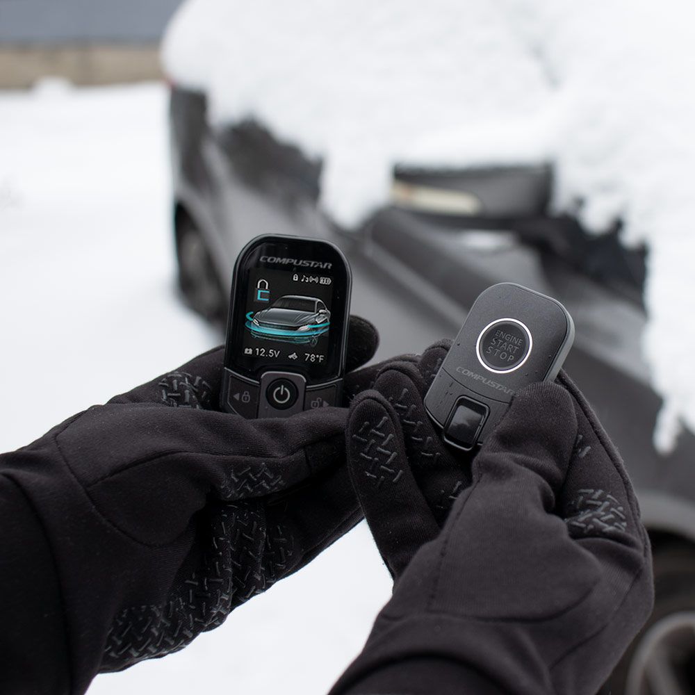Hands wearing black gloves holding a car key fob and remote. Snow-covered car in background.
