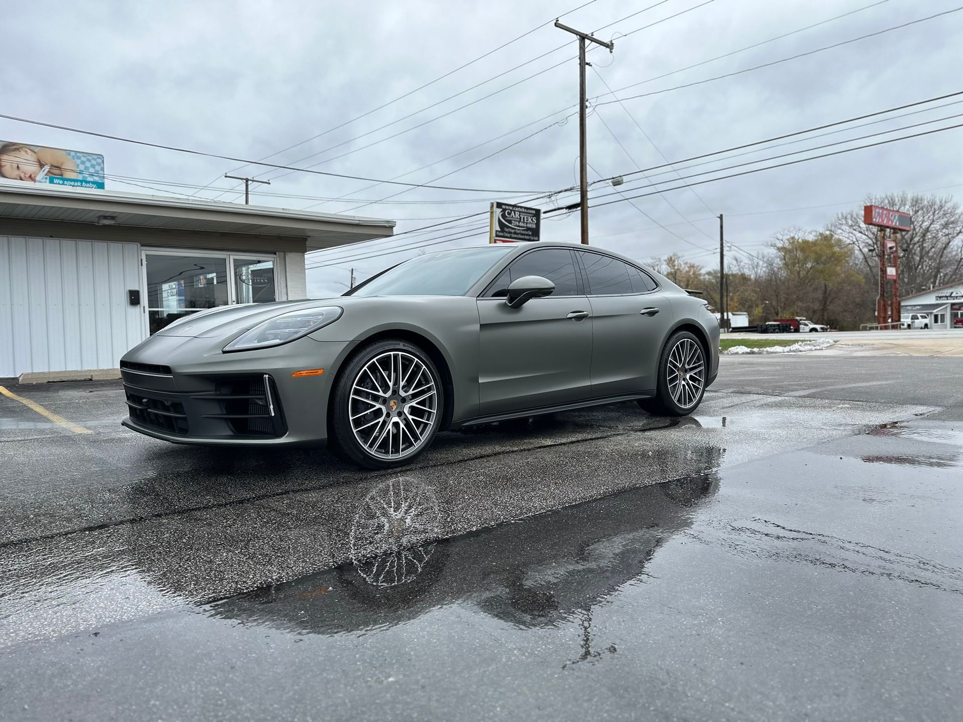 Gray Porsche Panamera parked on a wet street in front of a storefront on an overcast day.