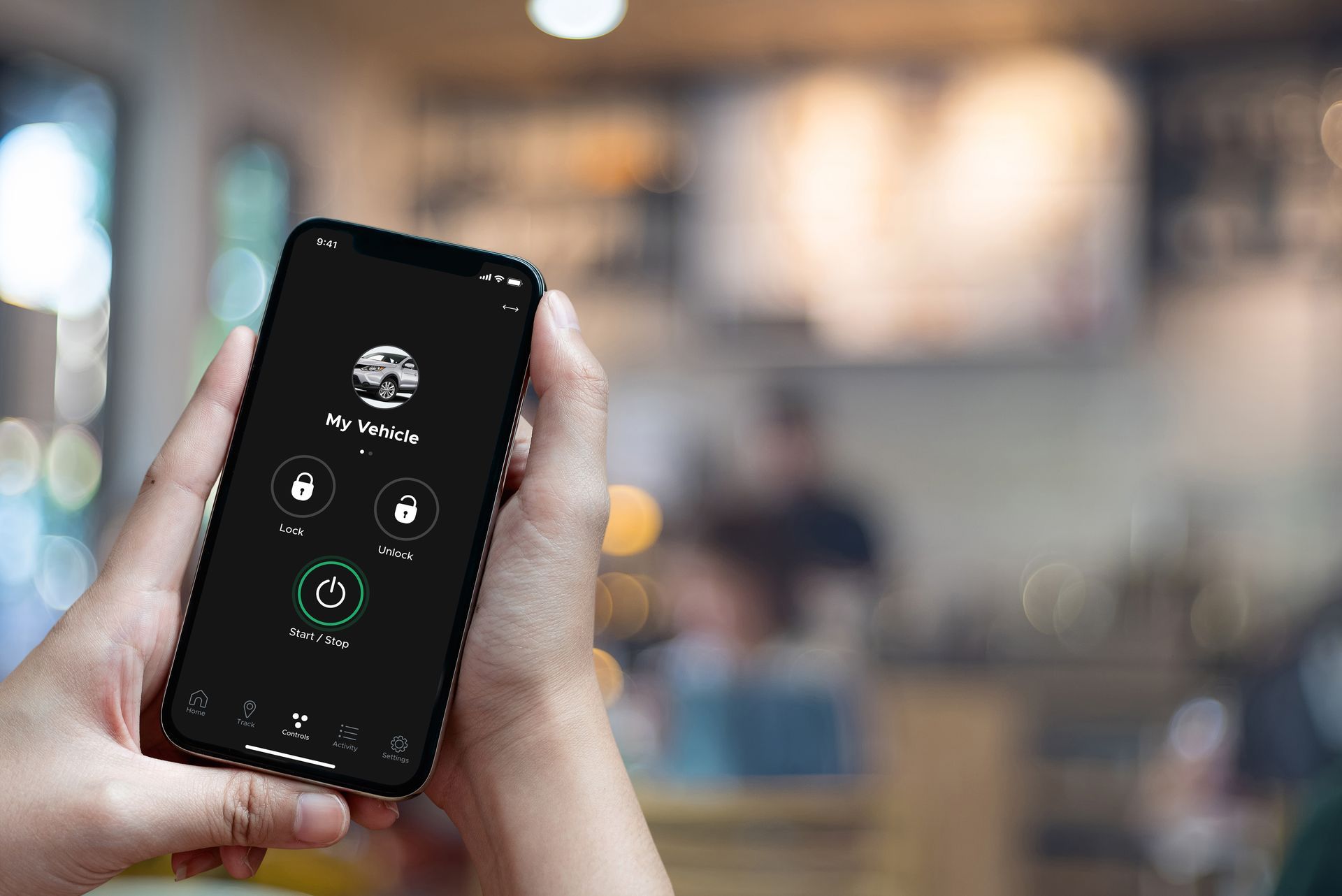 Hands holding a phone displaying a black app interface with vehicle control options, in a cafe setting.