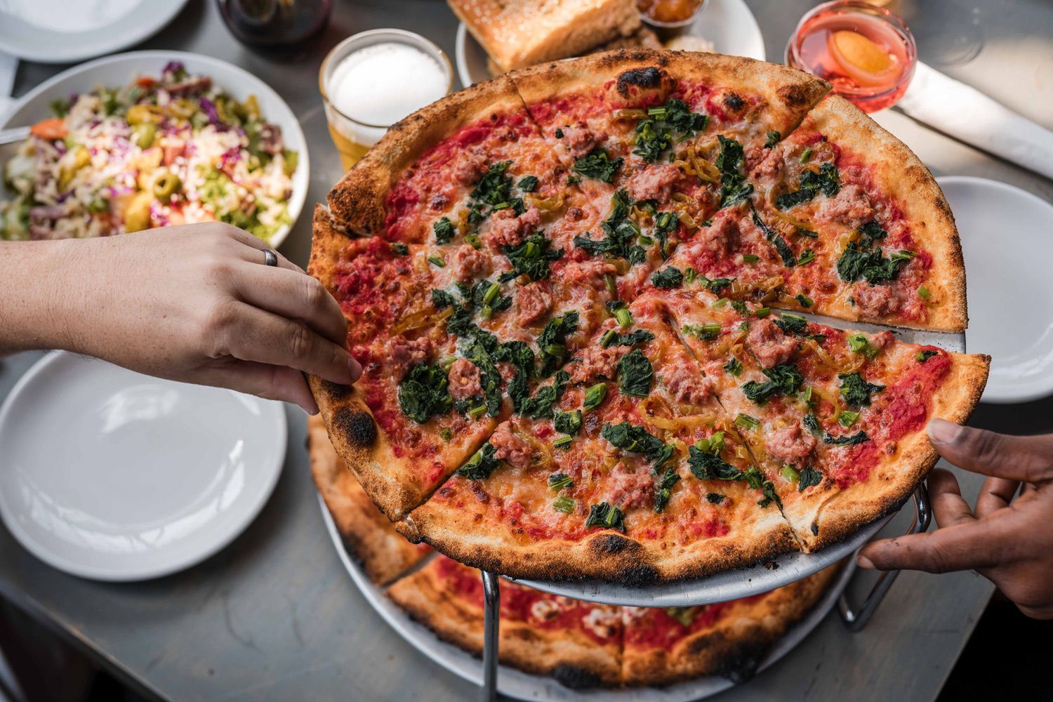 Freshly baked coal-fired pizza topped with Italian sausage, sautéed greens, and melted cheese at Nicky’s Coal Fired in Nashville, served alongside a colorful salad and drinks.