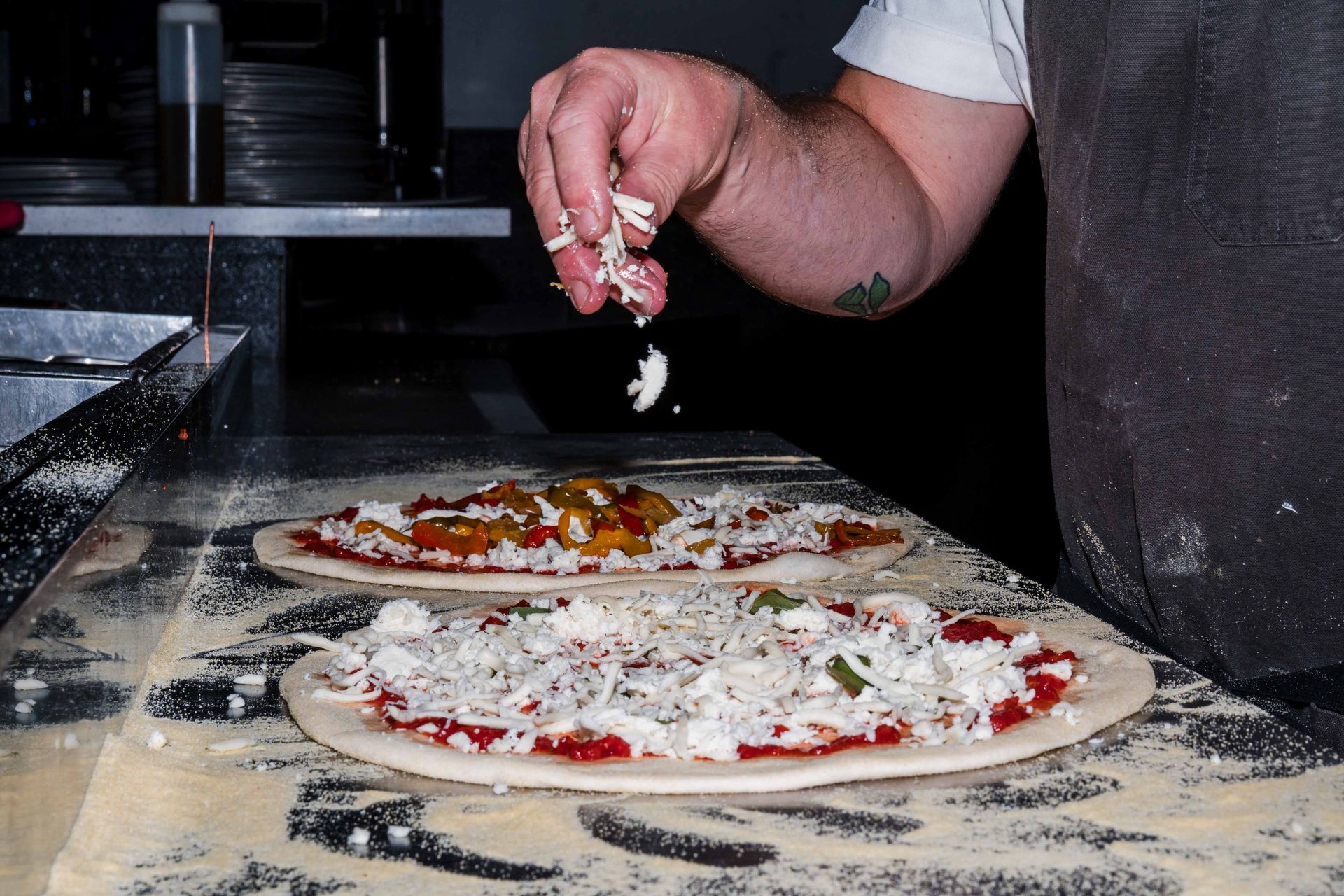 A close-up of a chef’s hand sprinkling fresh mozzarella cheese over raw pizza dough topped with toma