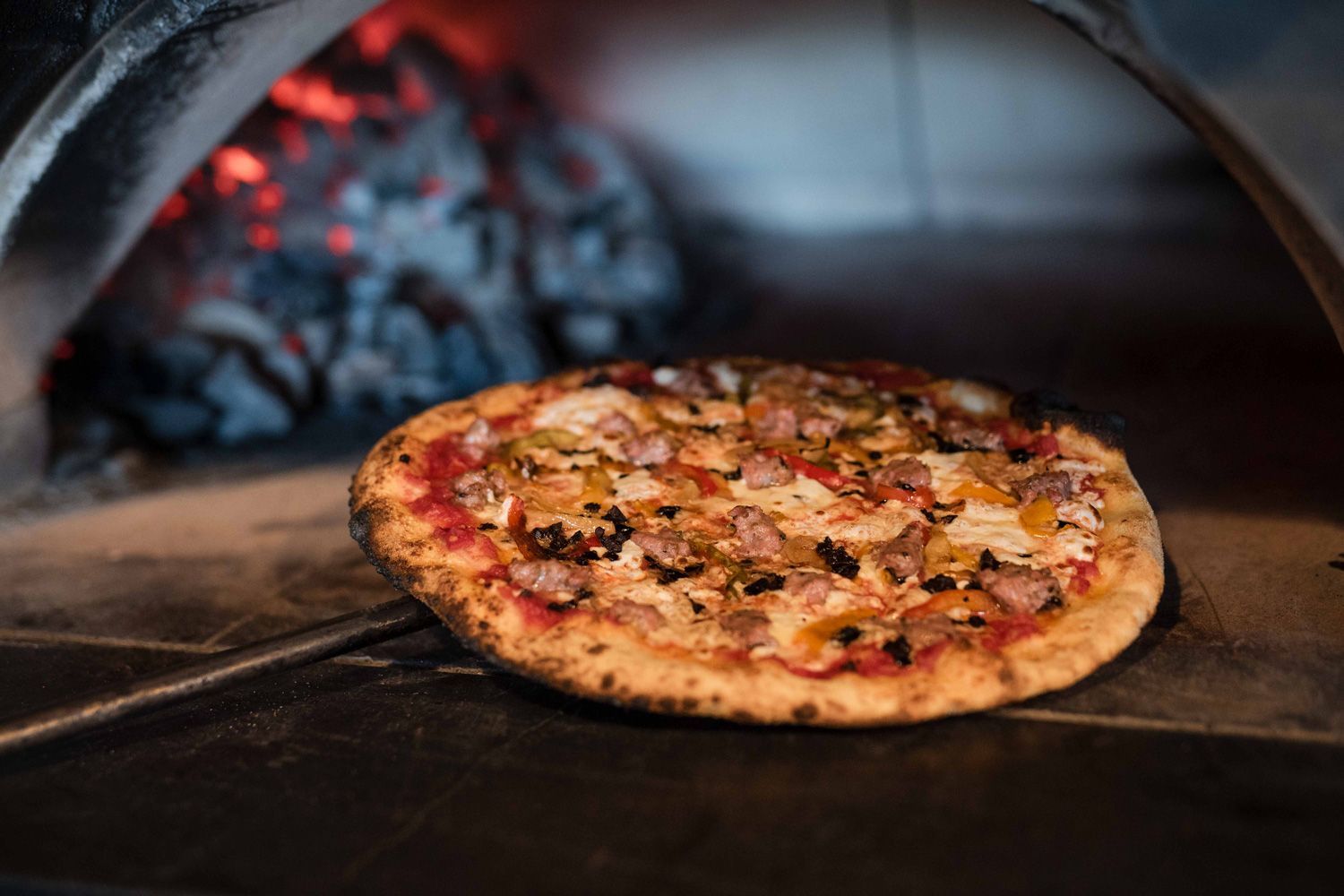 Coal-fired pizza being pulled from Nicky’s coal-fired oven, topped with sausage, peppers, and melted