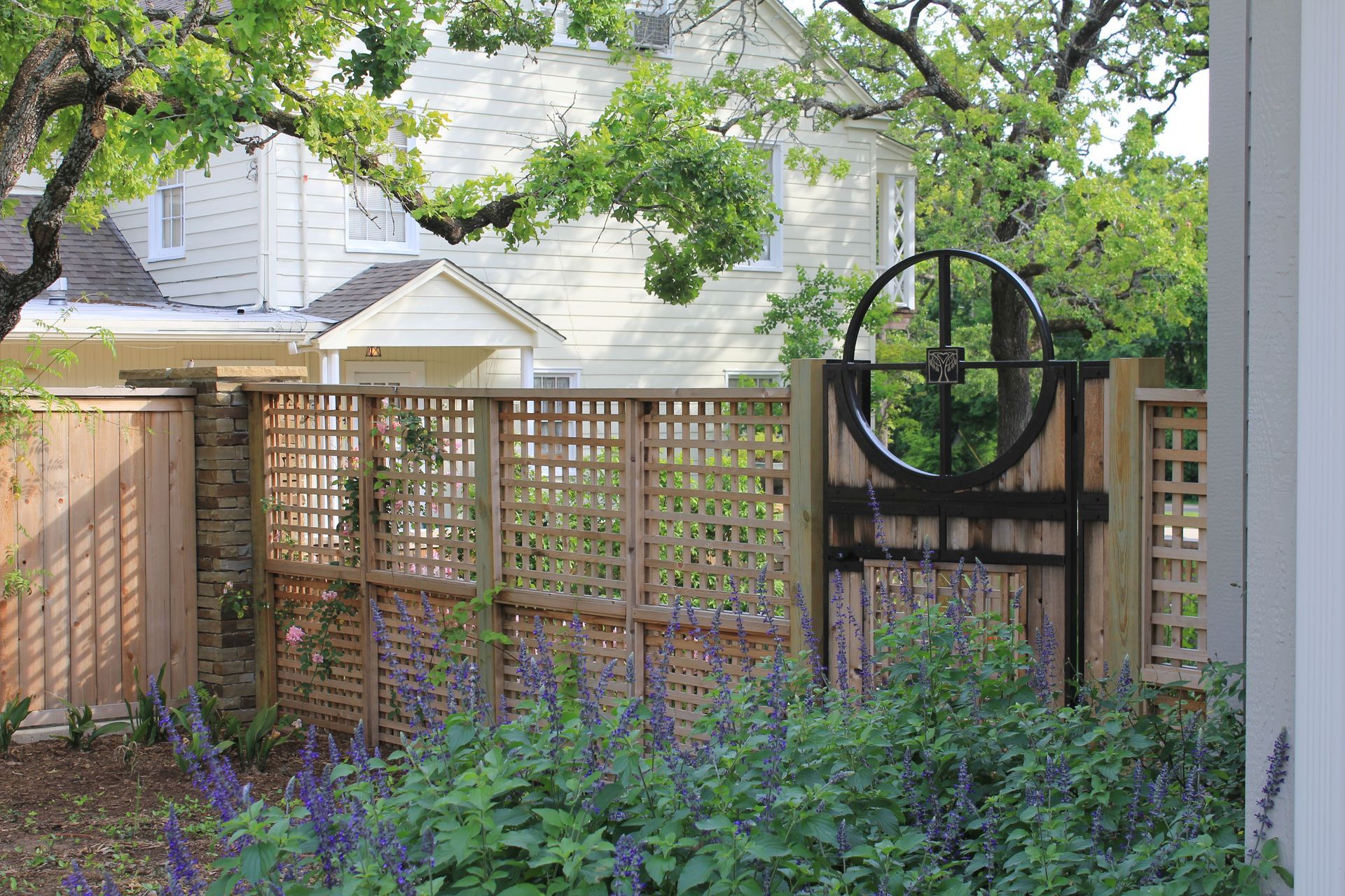 A wooden fence surrounds a garden with flowers and a house in the background.