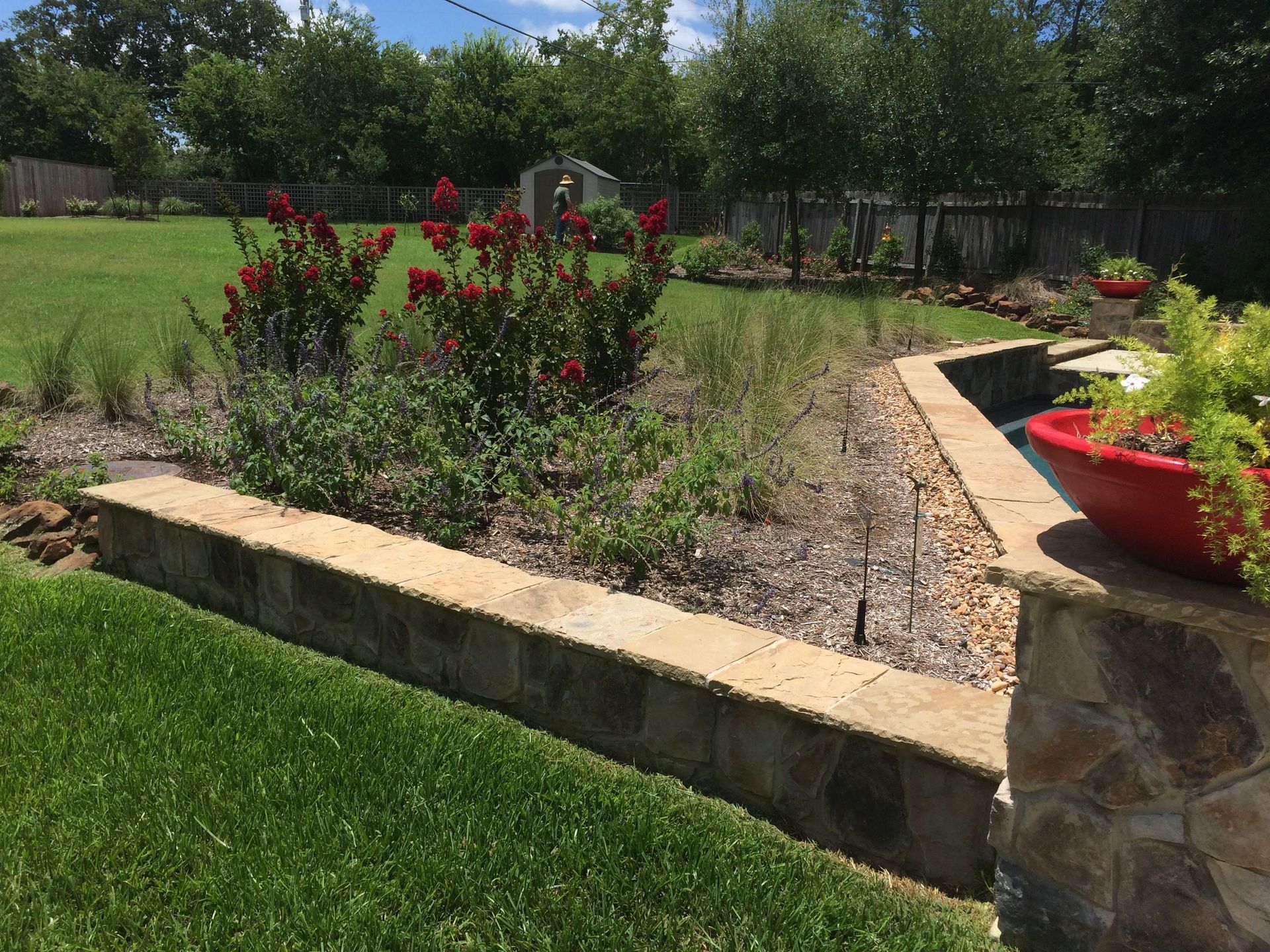 A lush green yard with a stone wall and a red potted plant.