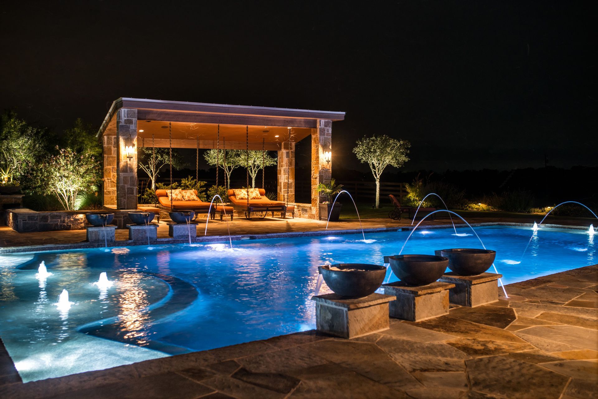 A large swimming pool is lit up at night with a gazebo in the background.