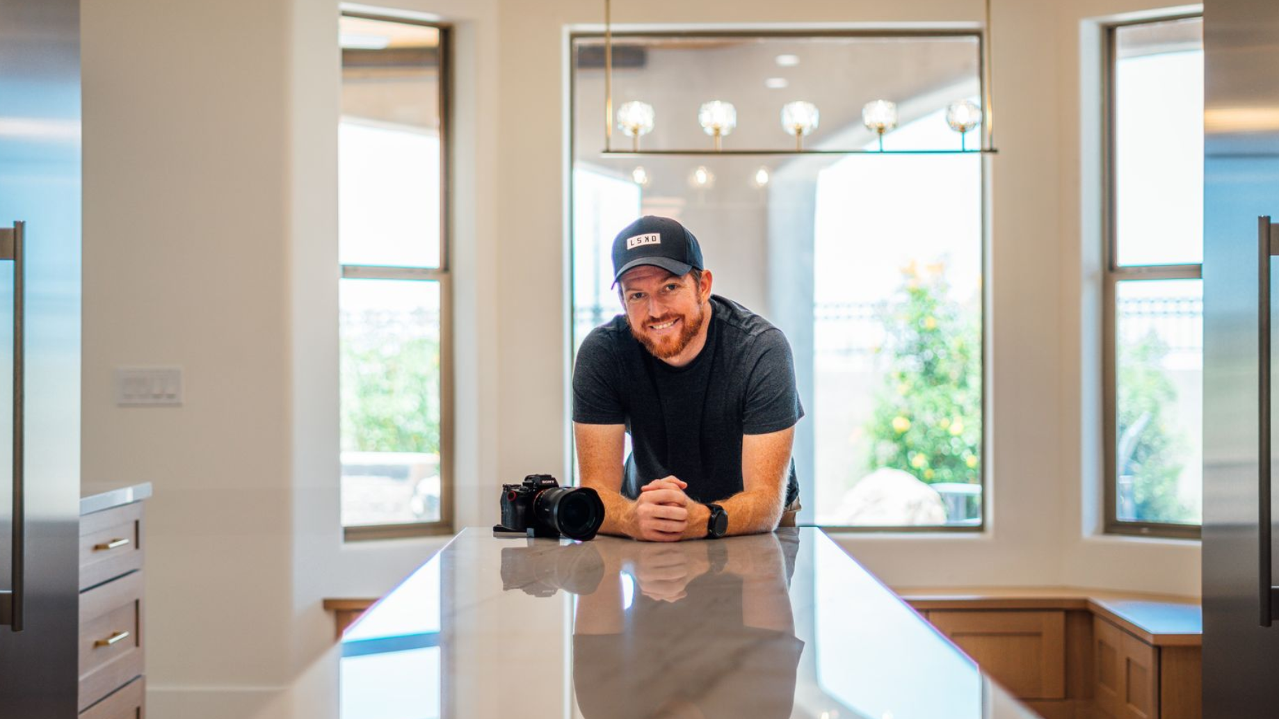 Man in a maroon shirt and gray cap sitting on a step in an empty, modern kitchen.