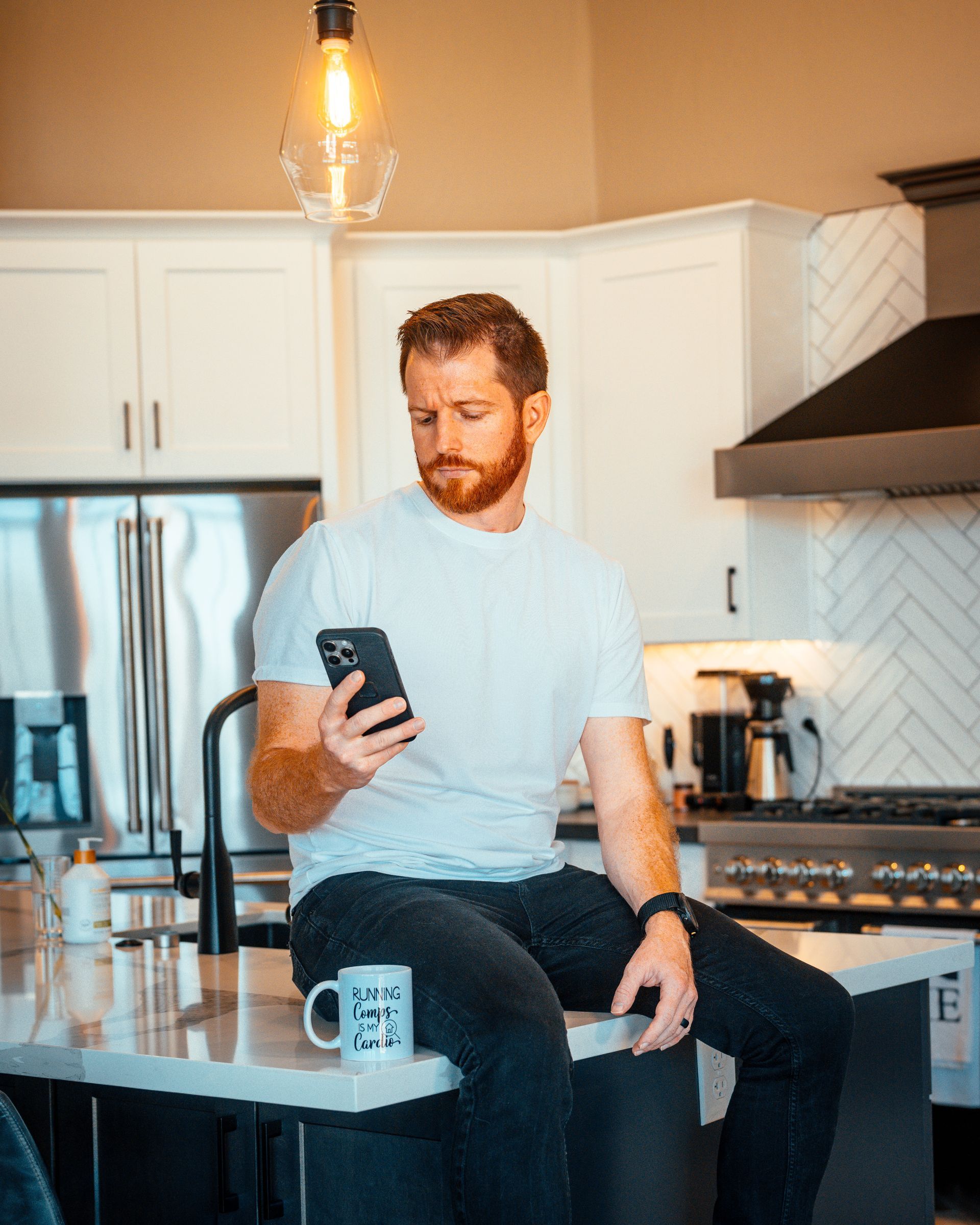 Man in white shirt looking at phone, sitting on kitchen countertop with coffee mug.