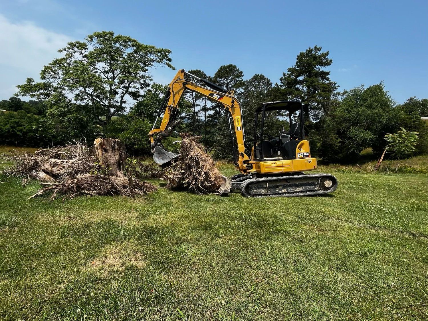 A yellow excavator is moving a pile of logs in a grassy field.